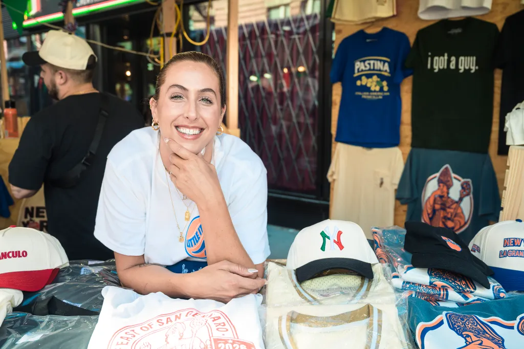 Nicole Ponti, of BODY, smiling at the camera, leaning on a table of merchandise from Red Sauce Studio during the Feast of San Gennaro in Little Italy.