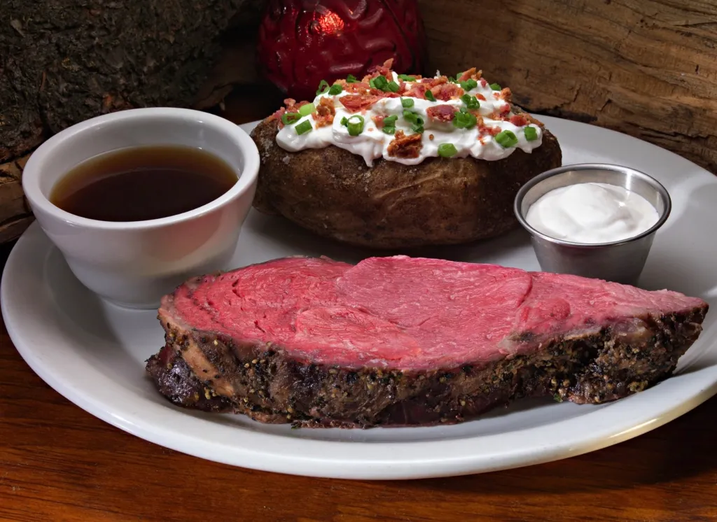 A plate of prime rib, a baked potato with toppings, and a cup of au jus.