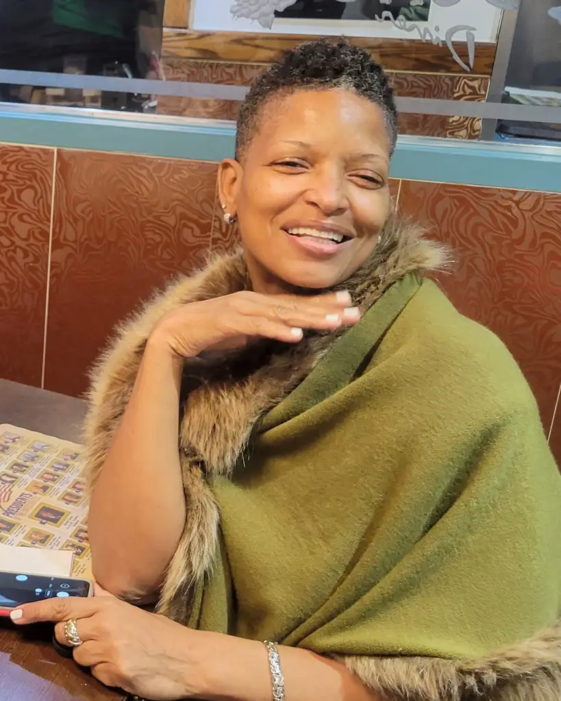 Alisa Goods, a Brooklyn grandmother, wearing a green shawl with a fur trim, smiling and touching her chin.