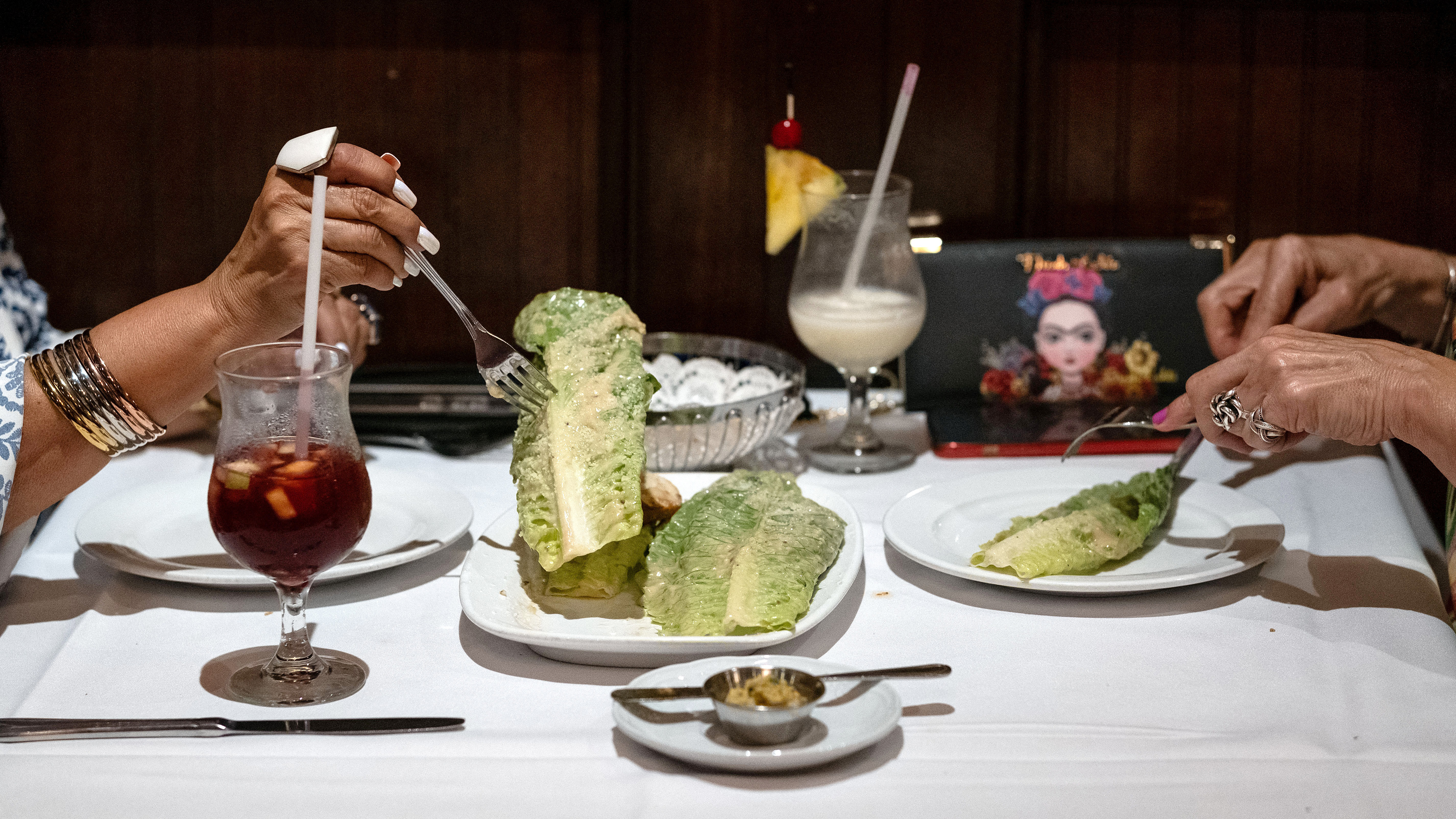 The hands of two people are visible using forks to pick up and cut gem lettuce at a restaurant table that also contains tropical drinks
