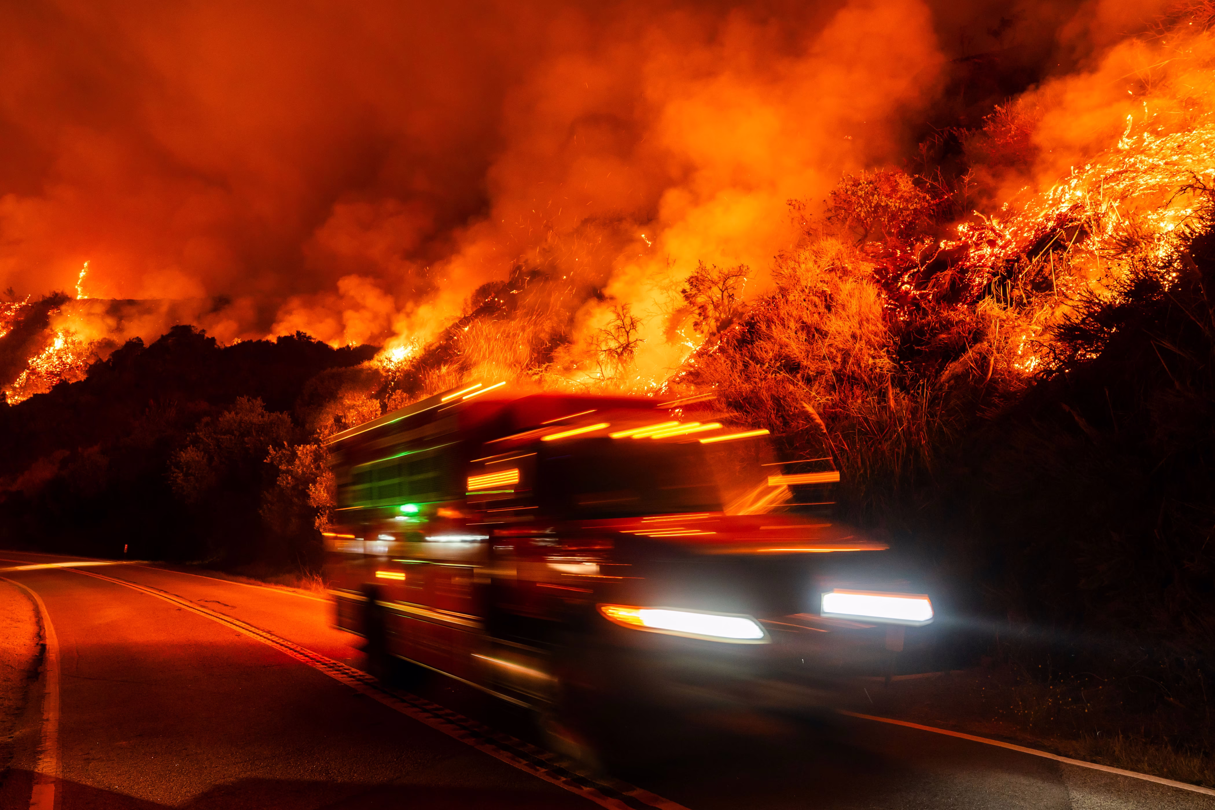 A firetruck passes on a road beneath a wildfire burning in the hills above, seen at night.