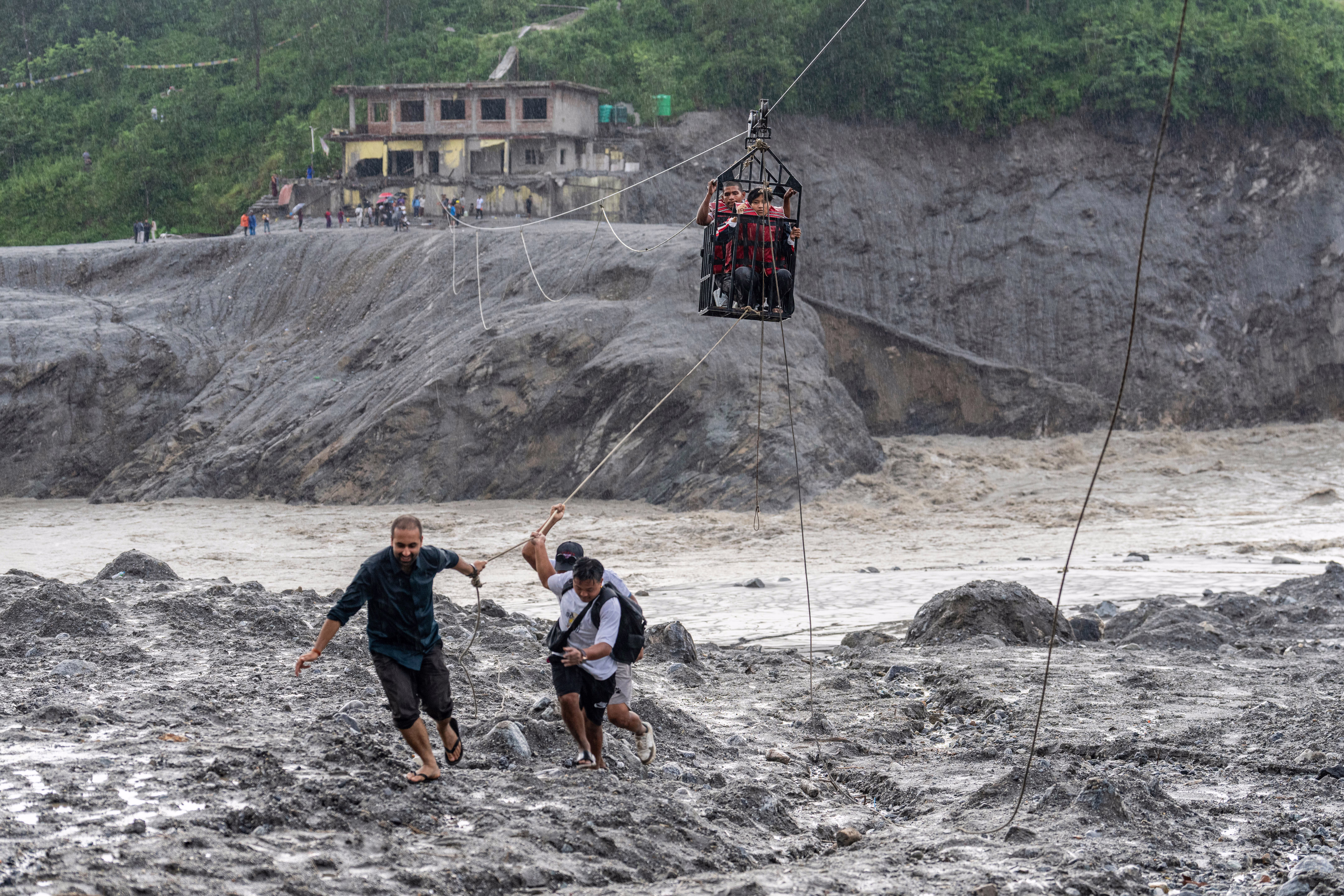 Several people on a riverbank pull a line attached to a basket hanging from a cable stretched across a river, helping people to cross.