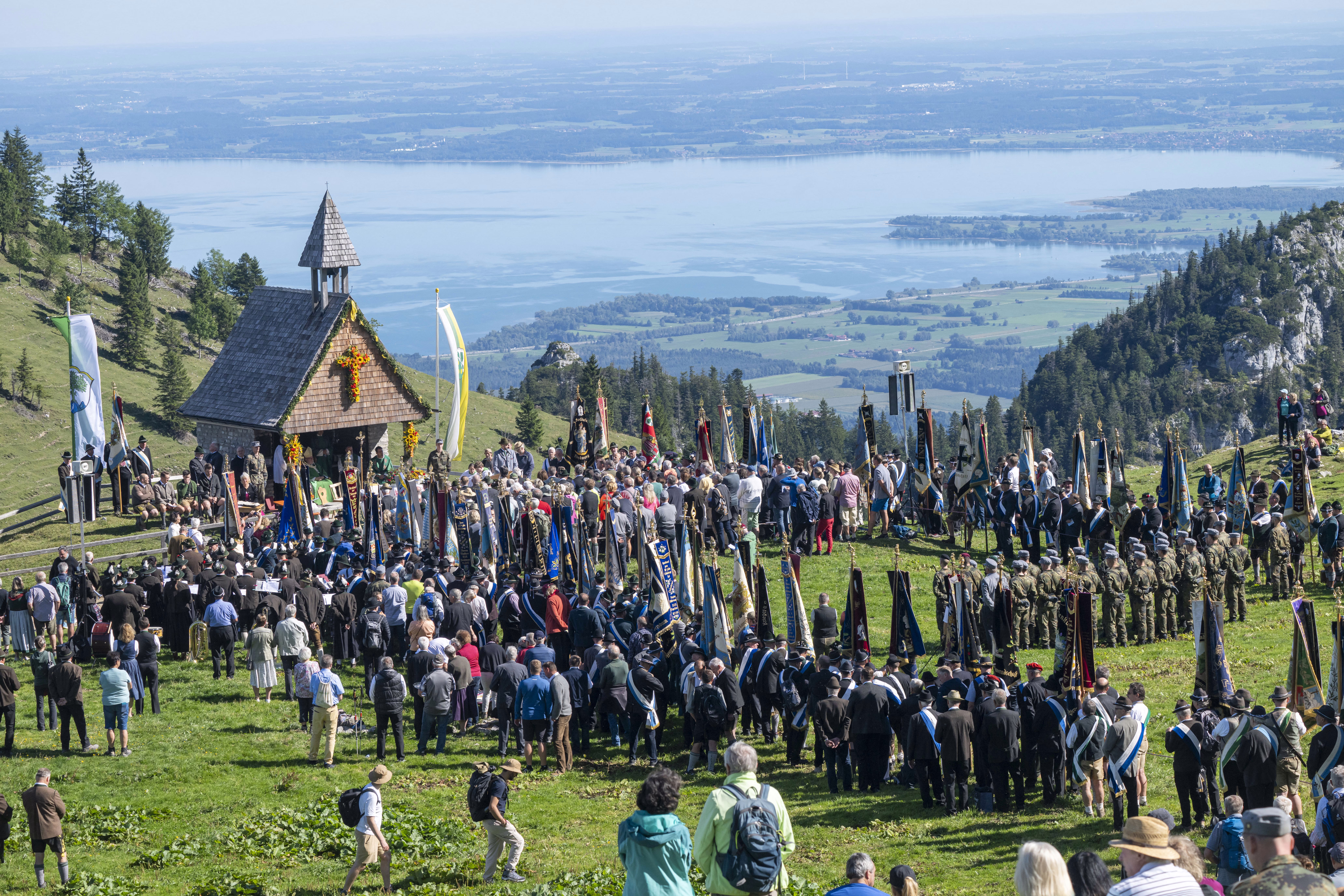 Hundreds of people attend a mountain Mass, standing in an Alpine meadow above a distant lake.