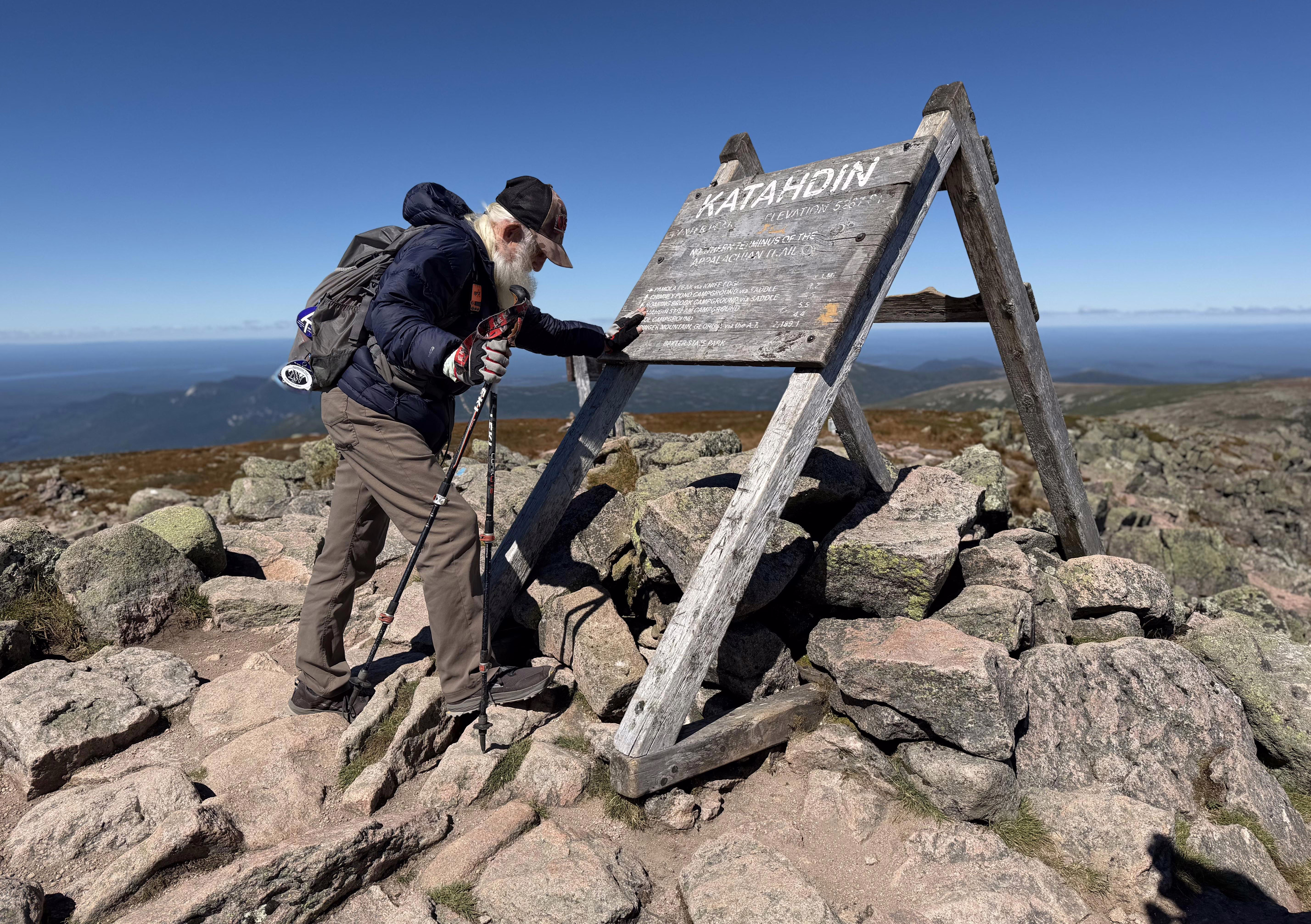 An older hiker pauses at the summit of a mountain, leaning a hand against a sign that reads 'Katahdin'.