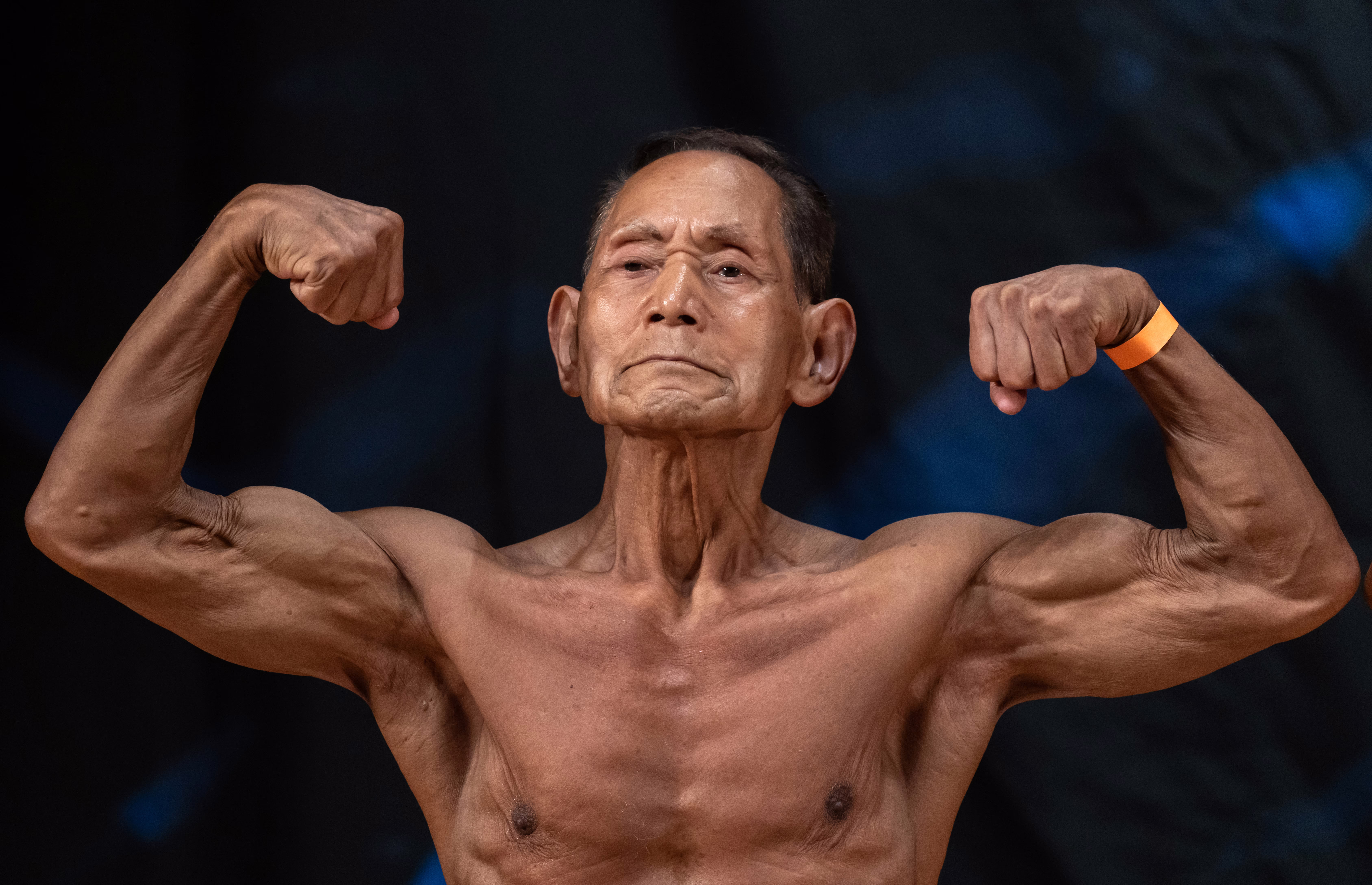 A 90-year-old bodybuilder flexes, arms raised, displaying impressive muscles.