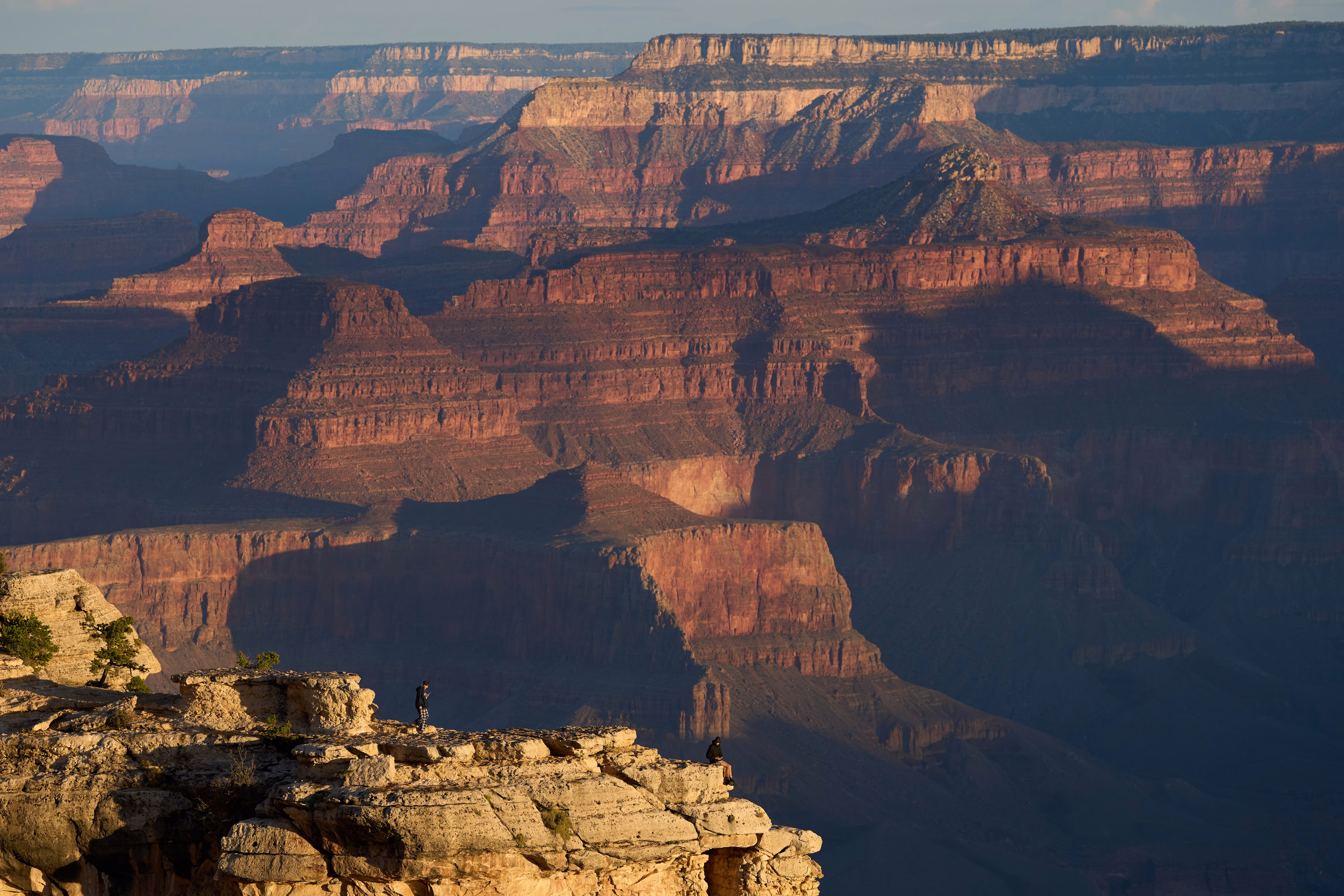 Two people stand and sit at the rim of the Grand Canyon, with a broad canyon vista beyond.
