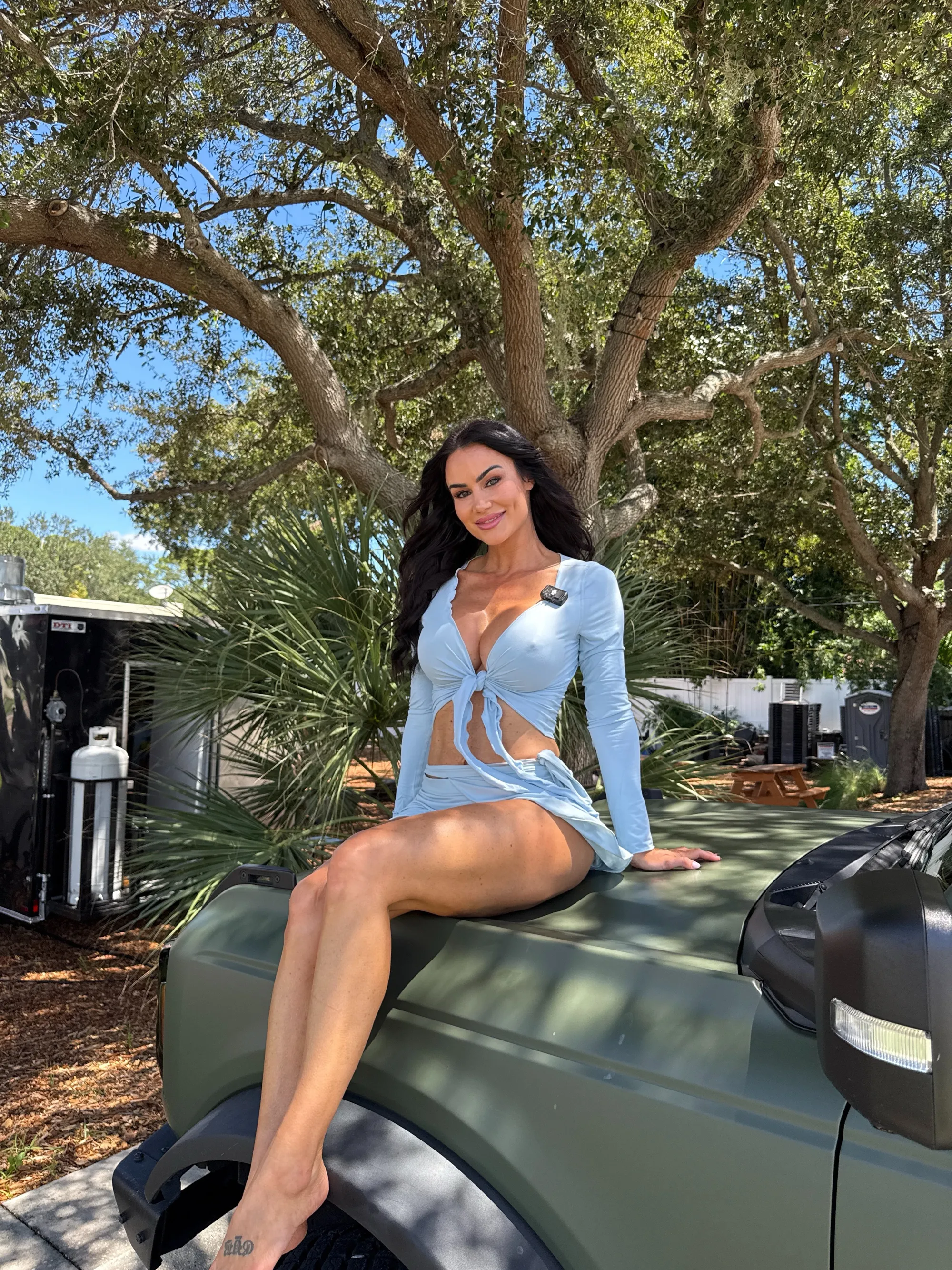 Catalina White, dressed in a light blue top and skirt, sitting on the hood of an olive green truck.