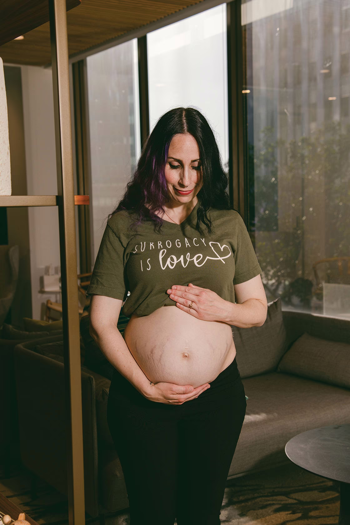 photo of woman with dark hair wearing t-shirt that reads 'SURROGACY IS LOVE' looks down lovingly at exposed pregnant belly