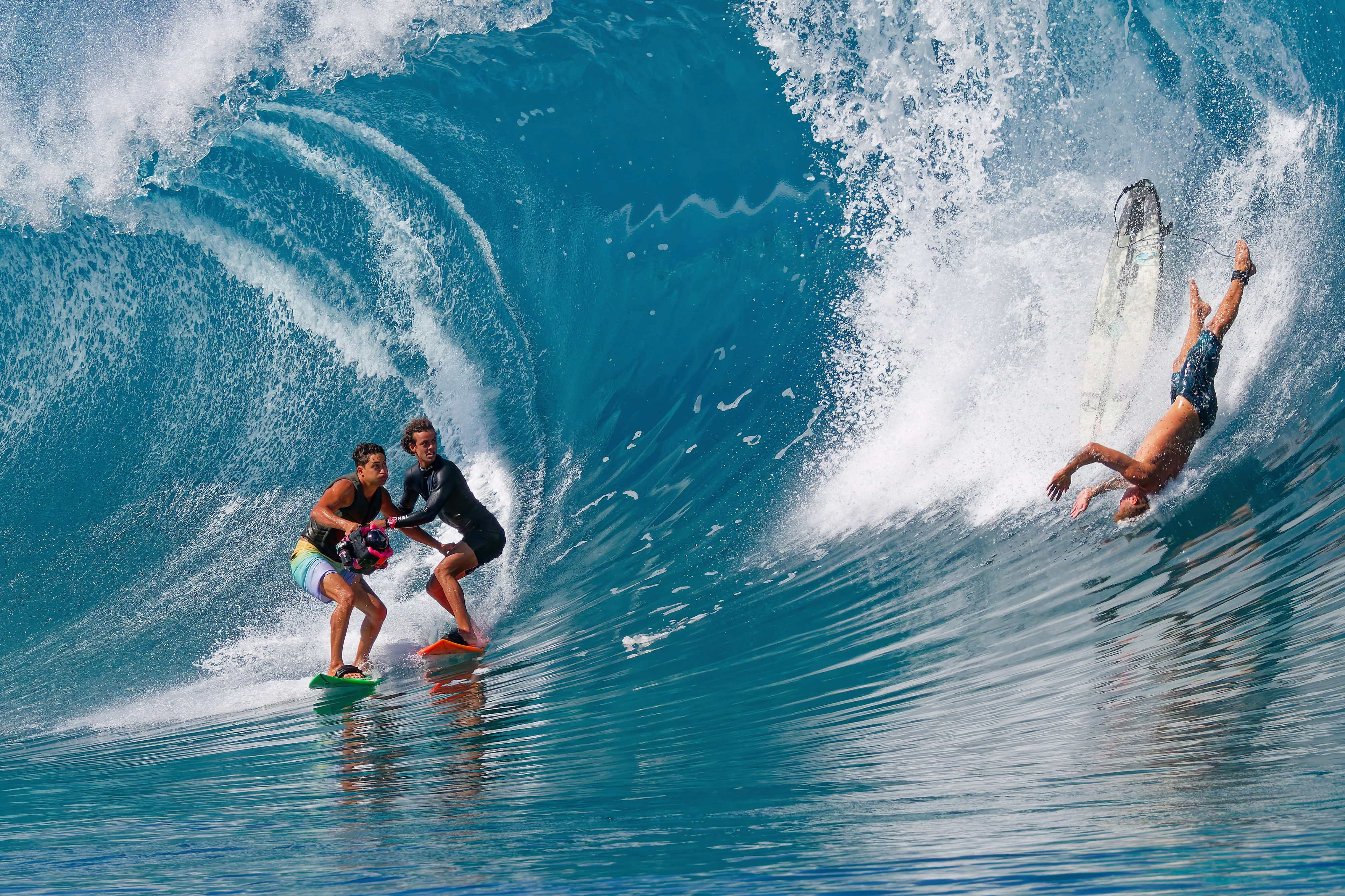 Three surfers on a very tall wave—two are standing, holding a camera between them, looking toward the third, who has fallen from their board and is tumbling upside-down just in front of the wave.