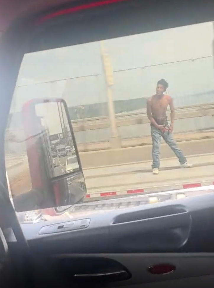 A shirtless man in jeans and white sneakers on a bridge, viewed through a car window.
