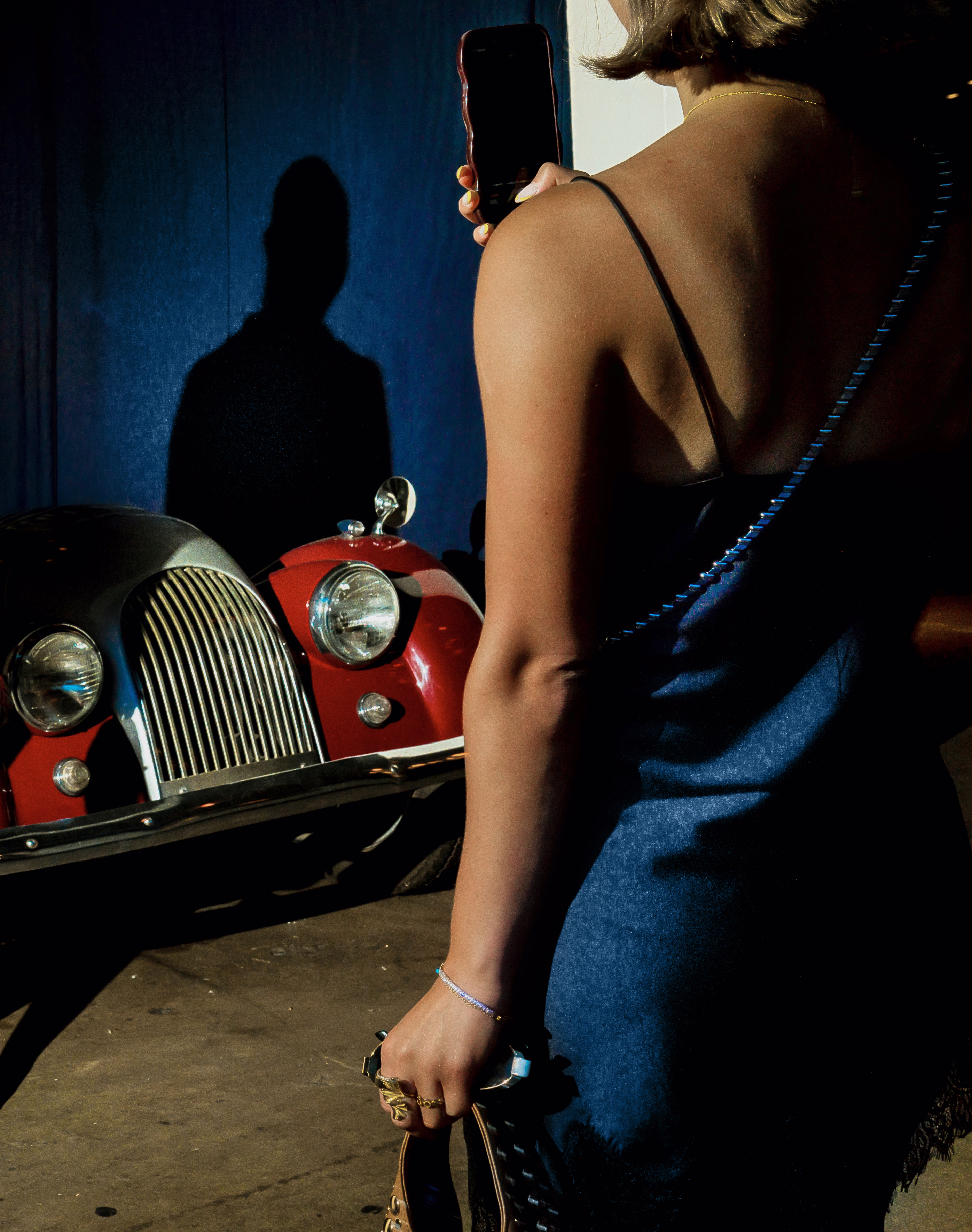 photo from behind of woman in blue spaghetti-strap dress taking cellphone photo of vintage red roadster with blue wall and man's shadow in background 