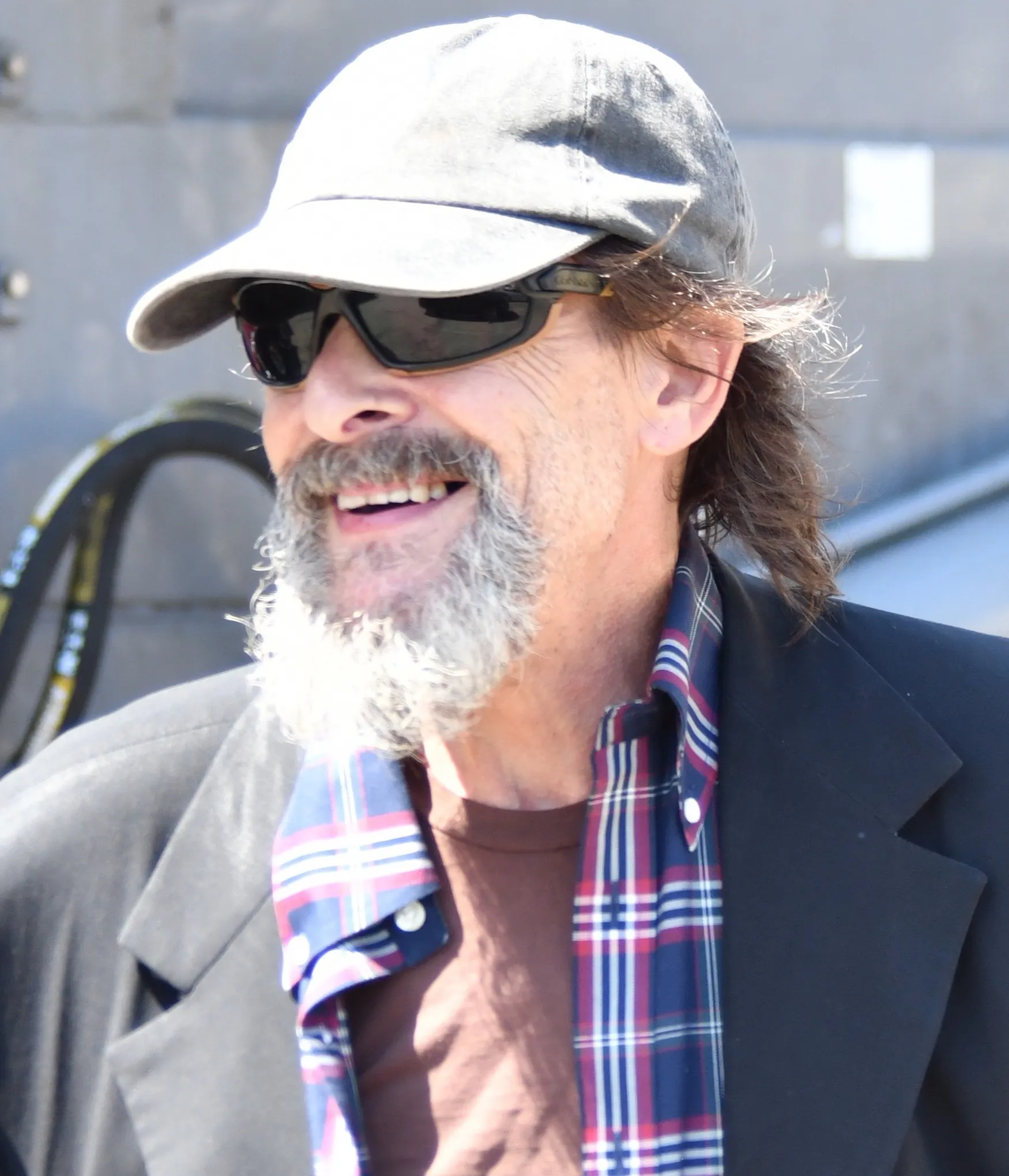 Judd Nelson smiling, wearing a baseball cap, sunglasses, and a plaid shirt under a black jacket.