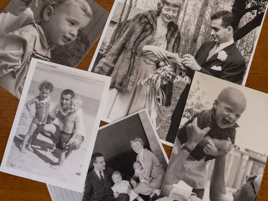 photo of an array of black-and-white family photos arranged on a wooden table, including portrait of child, two children at beach with man, wedding photo, family photo of couple with baby and toddler, and man lifting baby