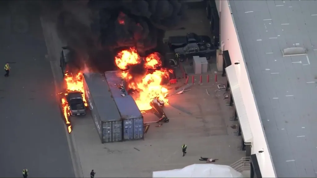 A helicopter crash with flames and dark smoke near shipping containers and a building.