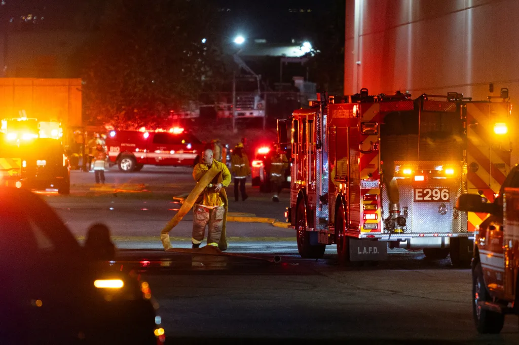 First responders at the scene of a helicopter crash in Chatsworth, Los Angeles, with a firefighter in yellow turnout gear holding a hose in the foreground and emergency vehicles with flashing lights in the background.