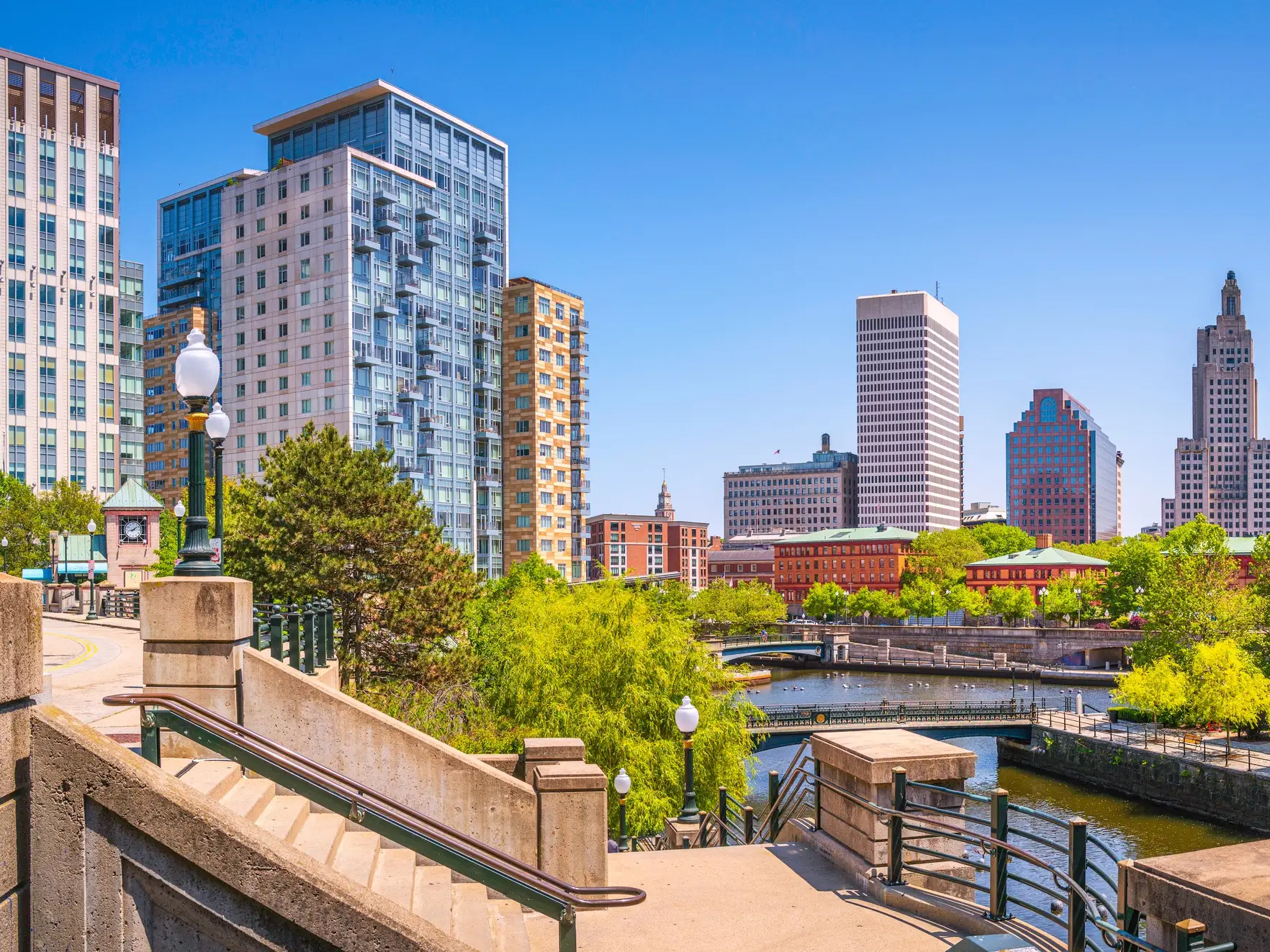 Downtown Providence skyline rises above riverwalk stairs, bridges, trees, and waterfront on a clear sunny day.