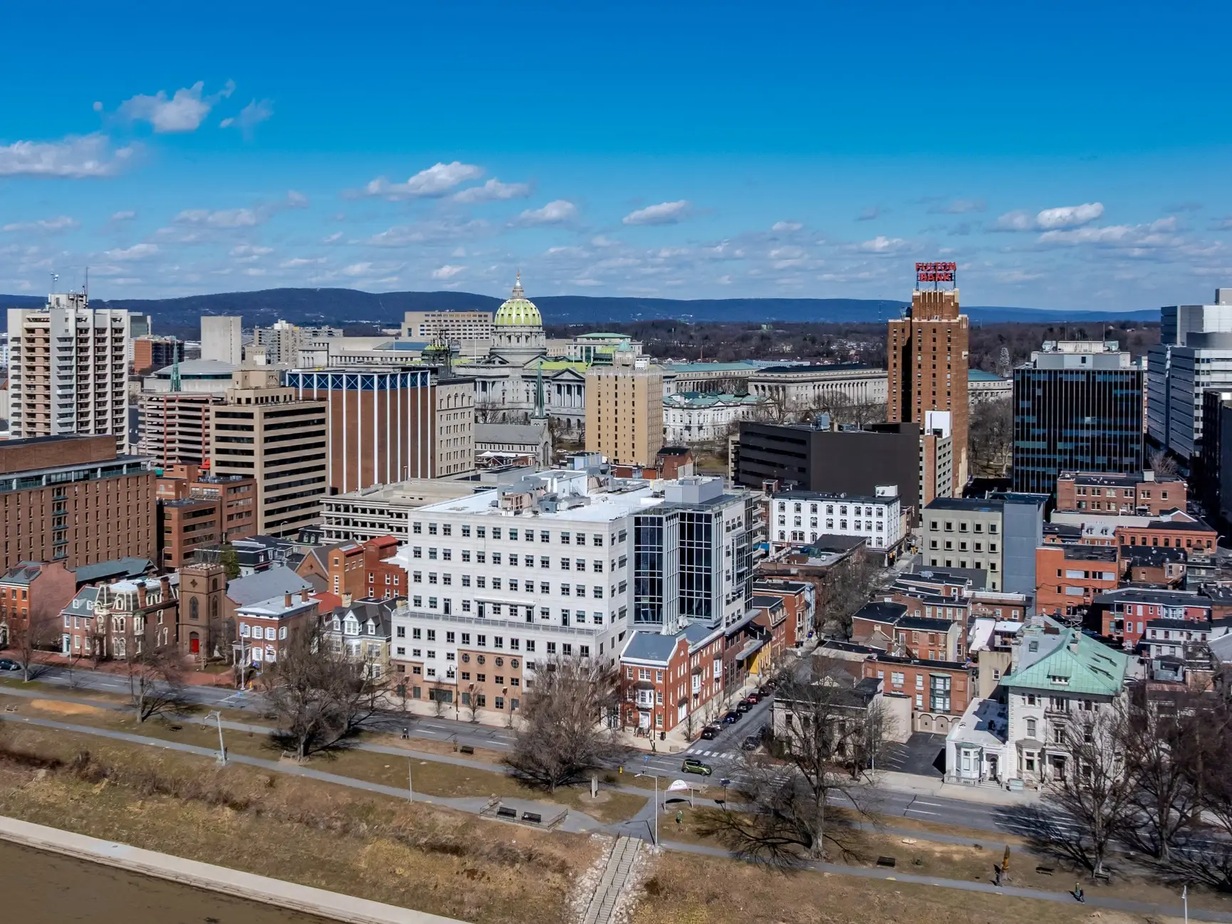 Downtown Harrisburg skyline with Pennsylvania State Capitol dome and riverfront buildings under a blue sky.