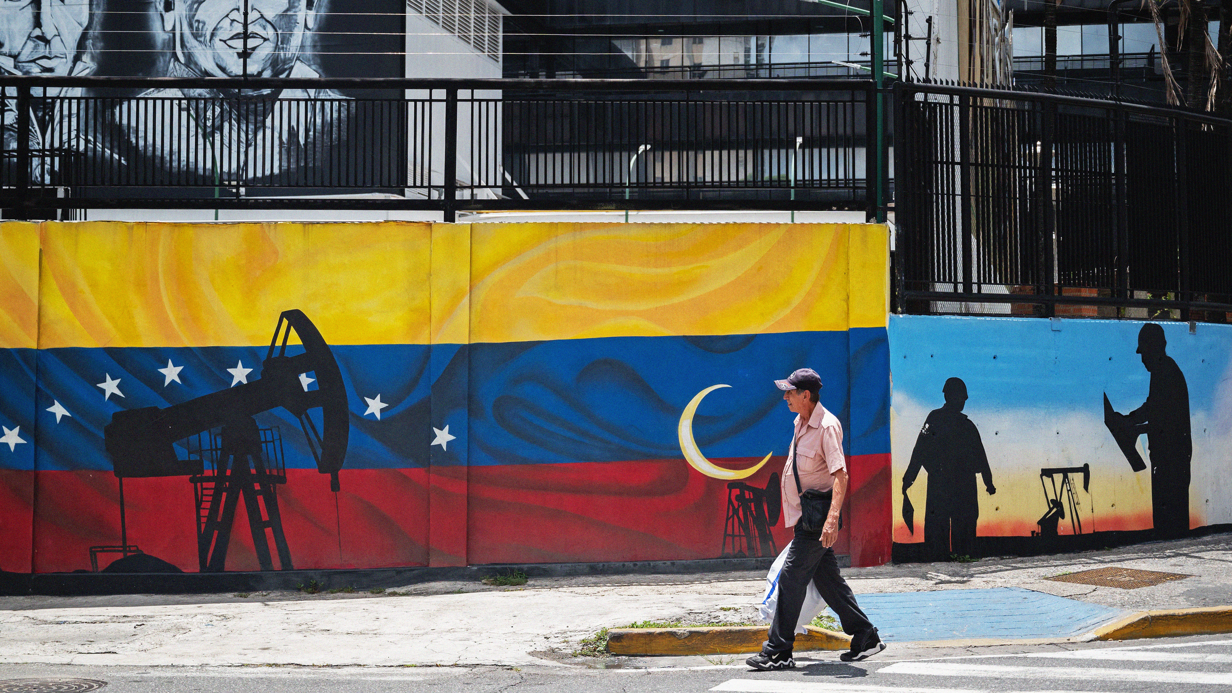 A man walks past a wall with a mural of an oil rig painted over a Venezuelan flag