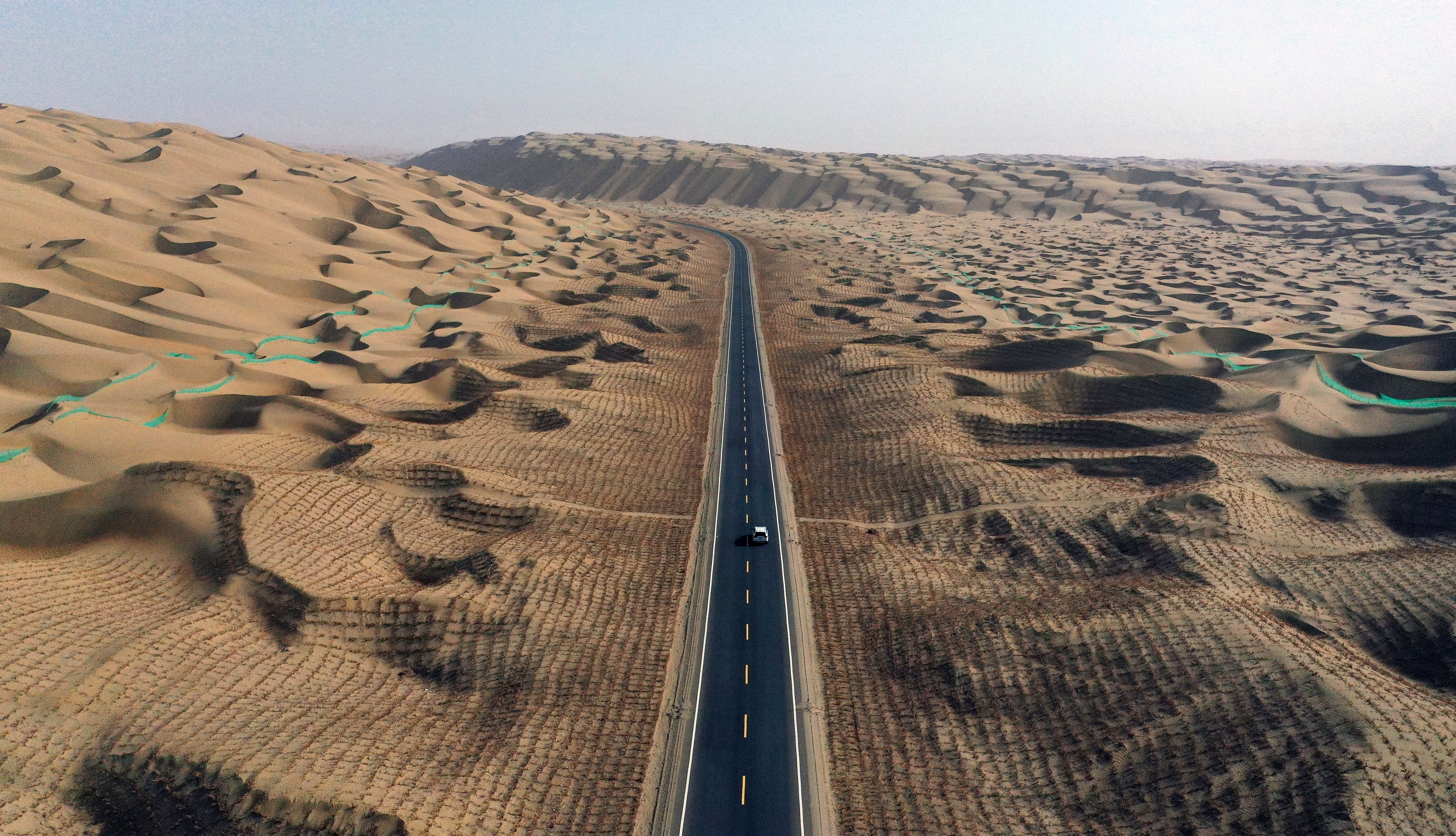 An aerial view of a highway in a desert, flanked by checkerboard-patterns of sand-control barriers on the dunes