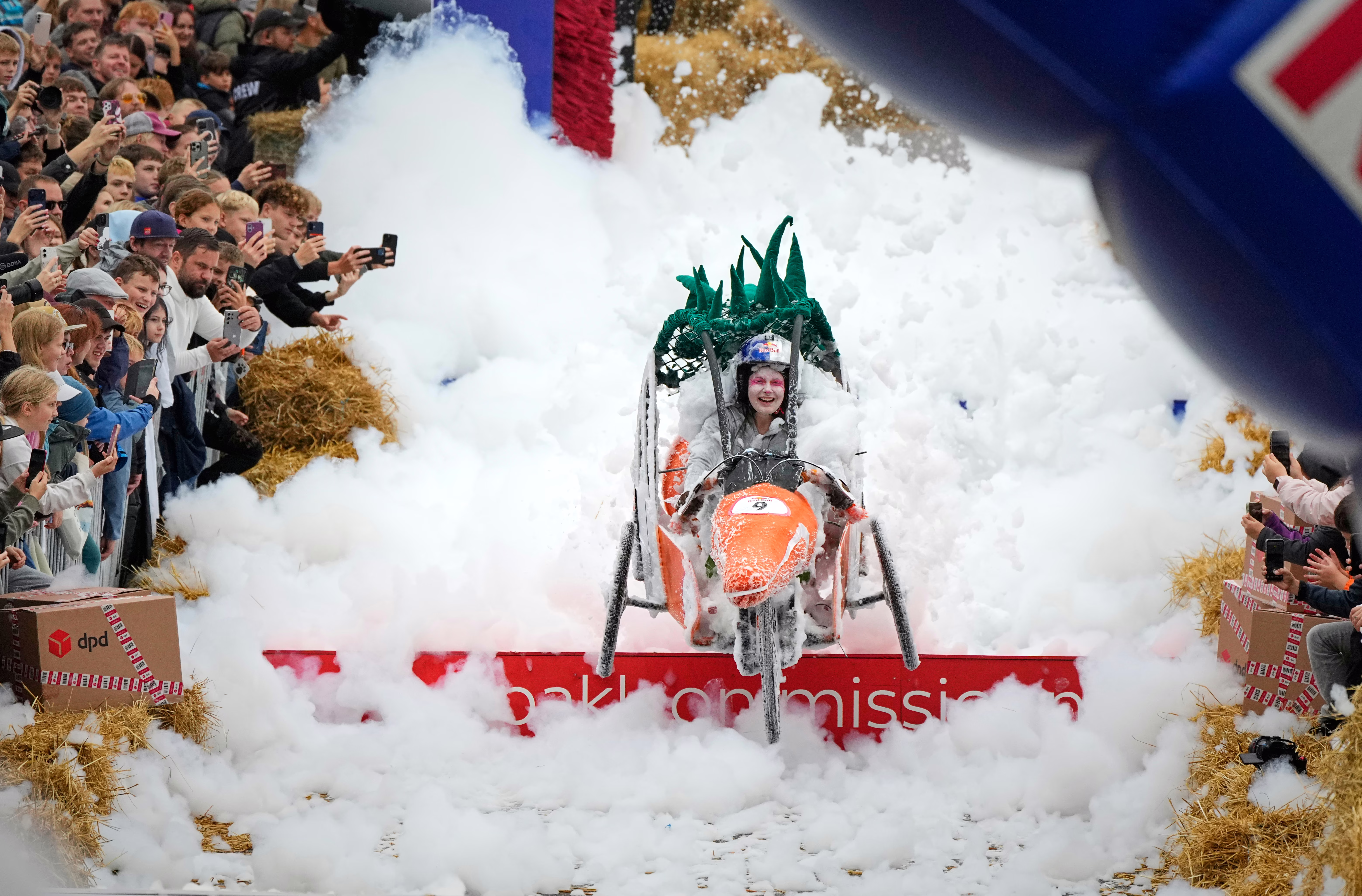 A crowd watches as a competitor in a small cart shaped like a carrot steers through a sudsy stretch in a soapbox obstacle course.