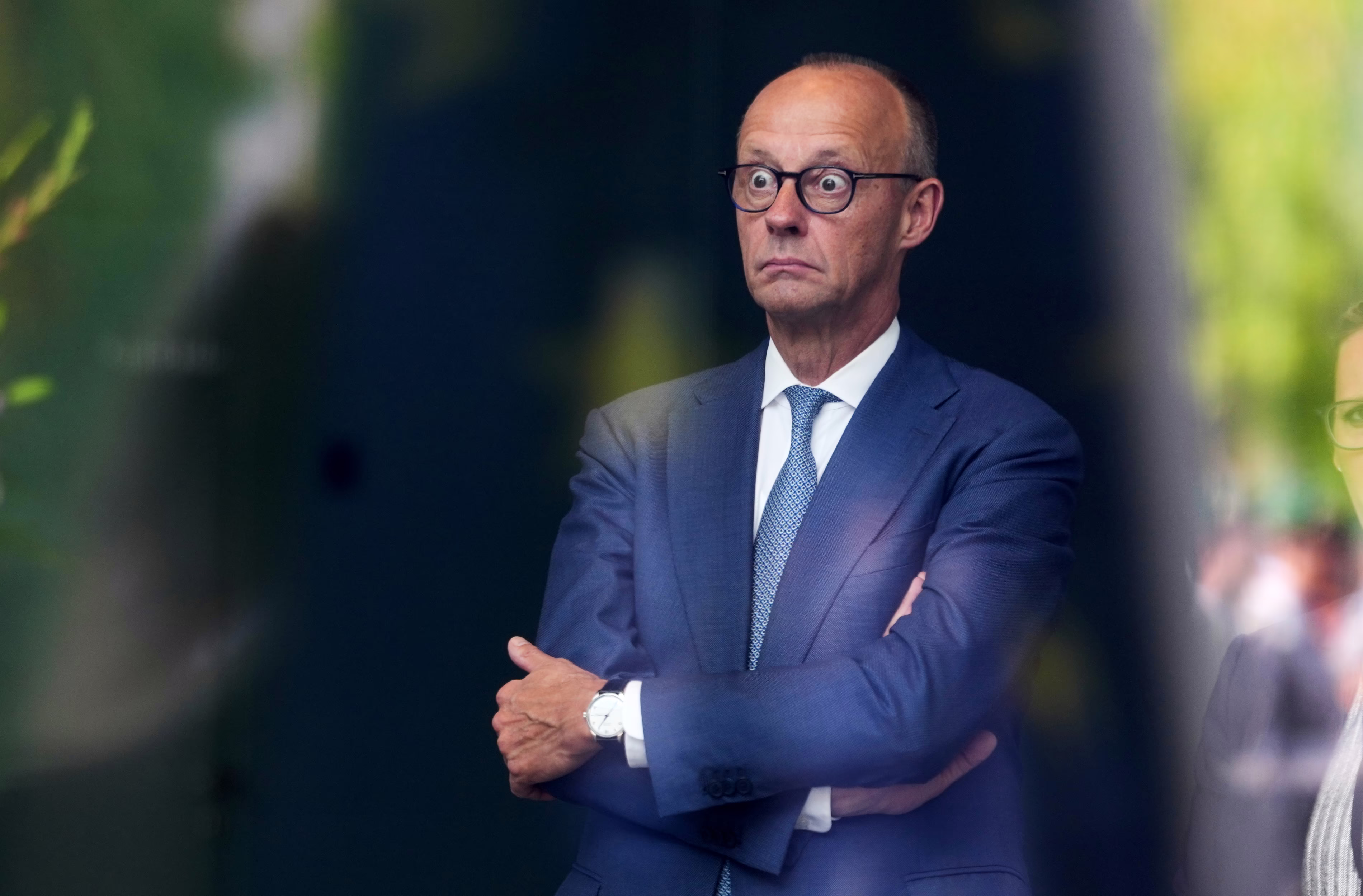 German Chancellor Friedrich Merz stands, arms crossed, eyes open very wide.