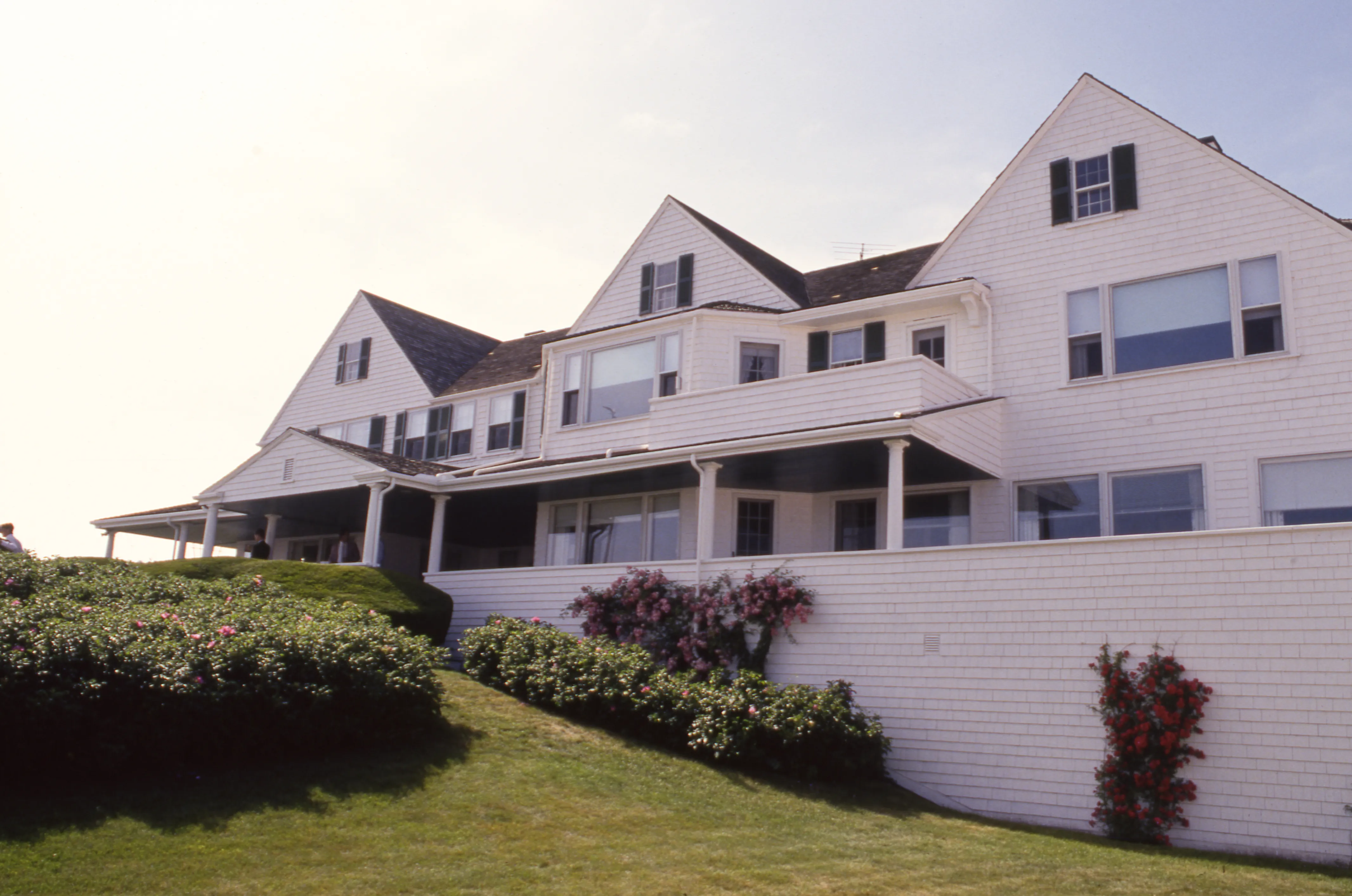 The residence of Senator Ted Kennedy in Hyannis Port, Massachusetts, featuring a white house with dark shutters and a large porch, surrounded by green shrubs and a grassy lawn.