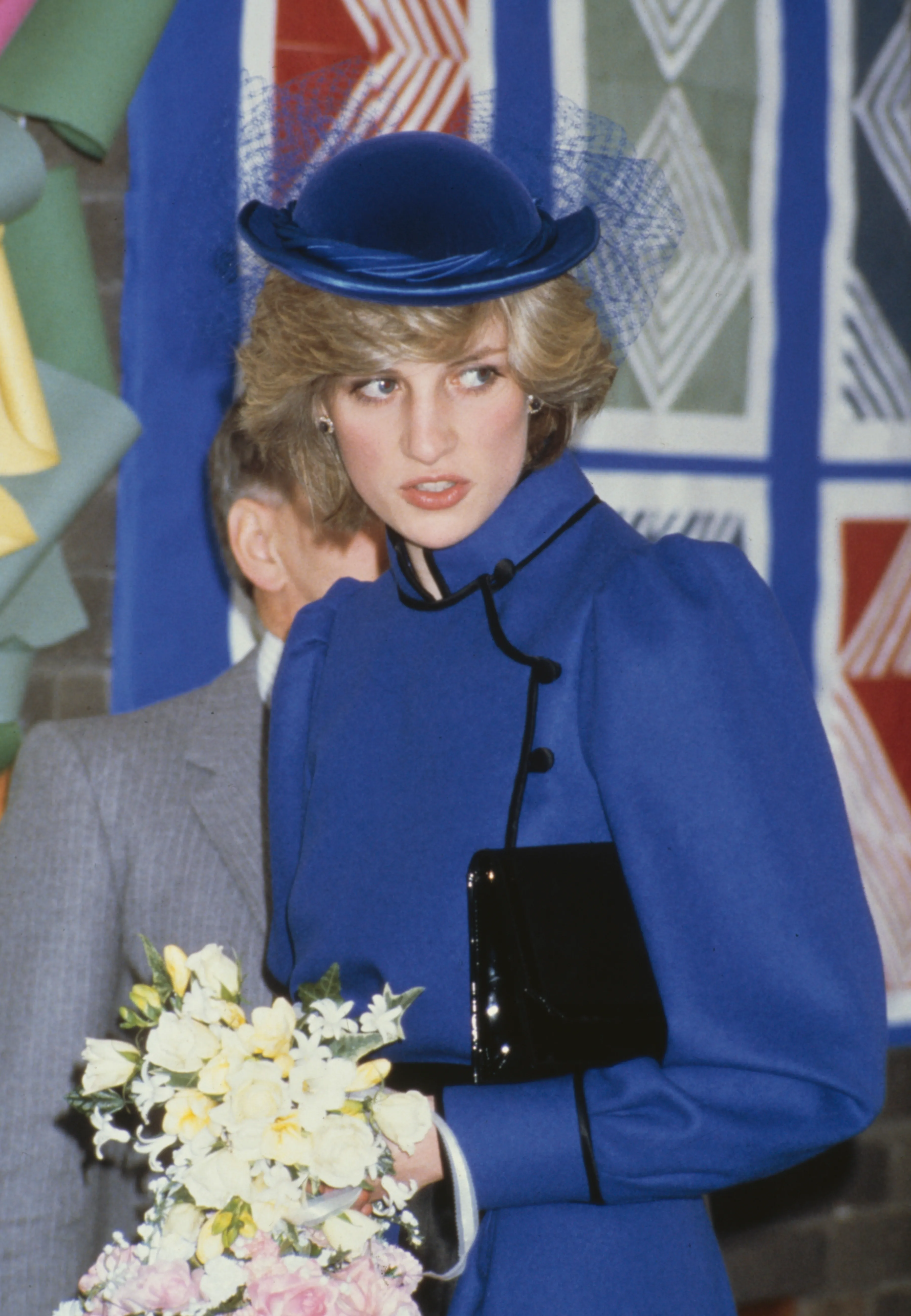 Diana, Princess of Wales, in a blue coat with black trim and a matching hat, carrying a bouquet of flowers.