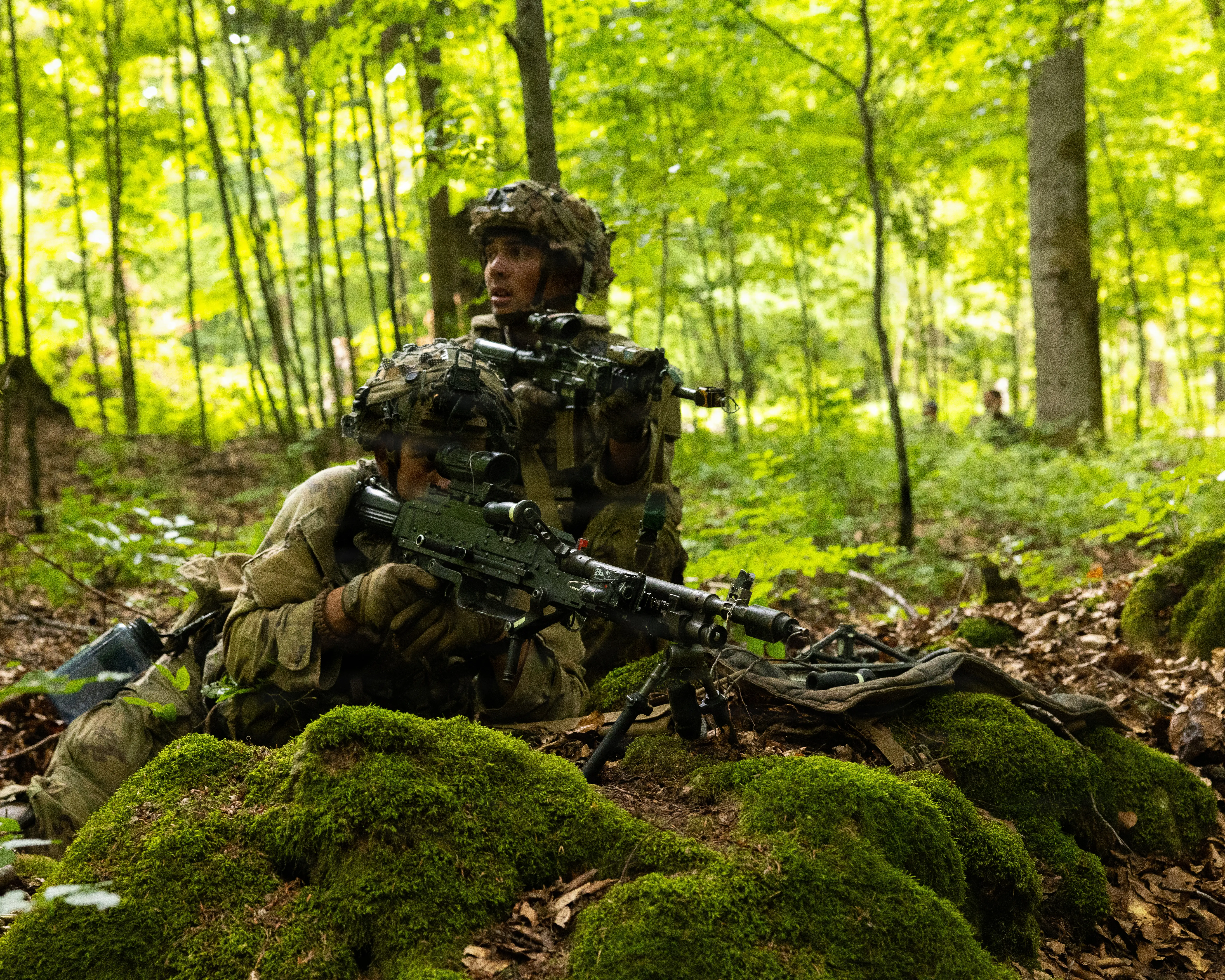 Two US Army soldiers crouch in position with rifles in the forest.