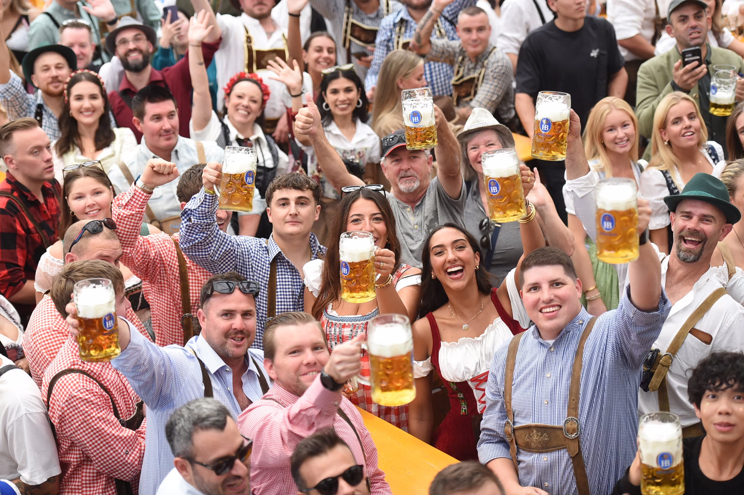 A crowd of people at Oktoberfest smile and raise mugs of beer toward the camera.
