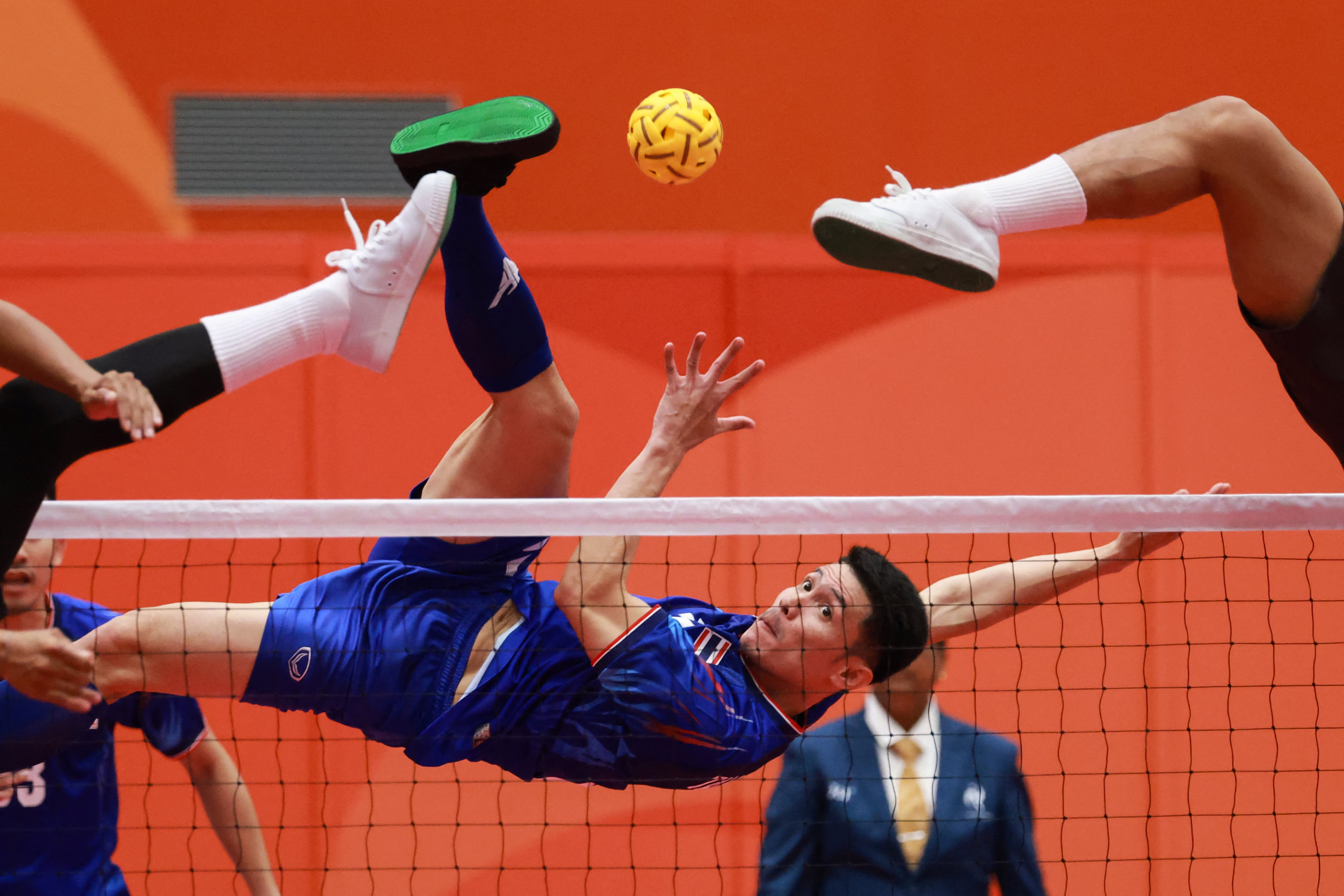 An athlete leaps high in the air to kick a small ball over a net, as defenders are seen with their feet also high in the air on the opposite side of the net.