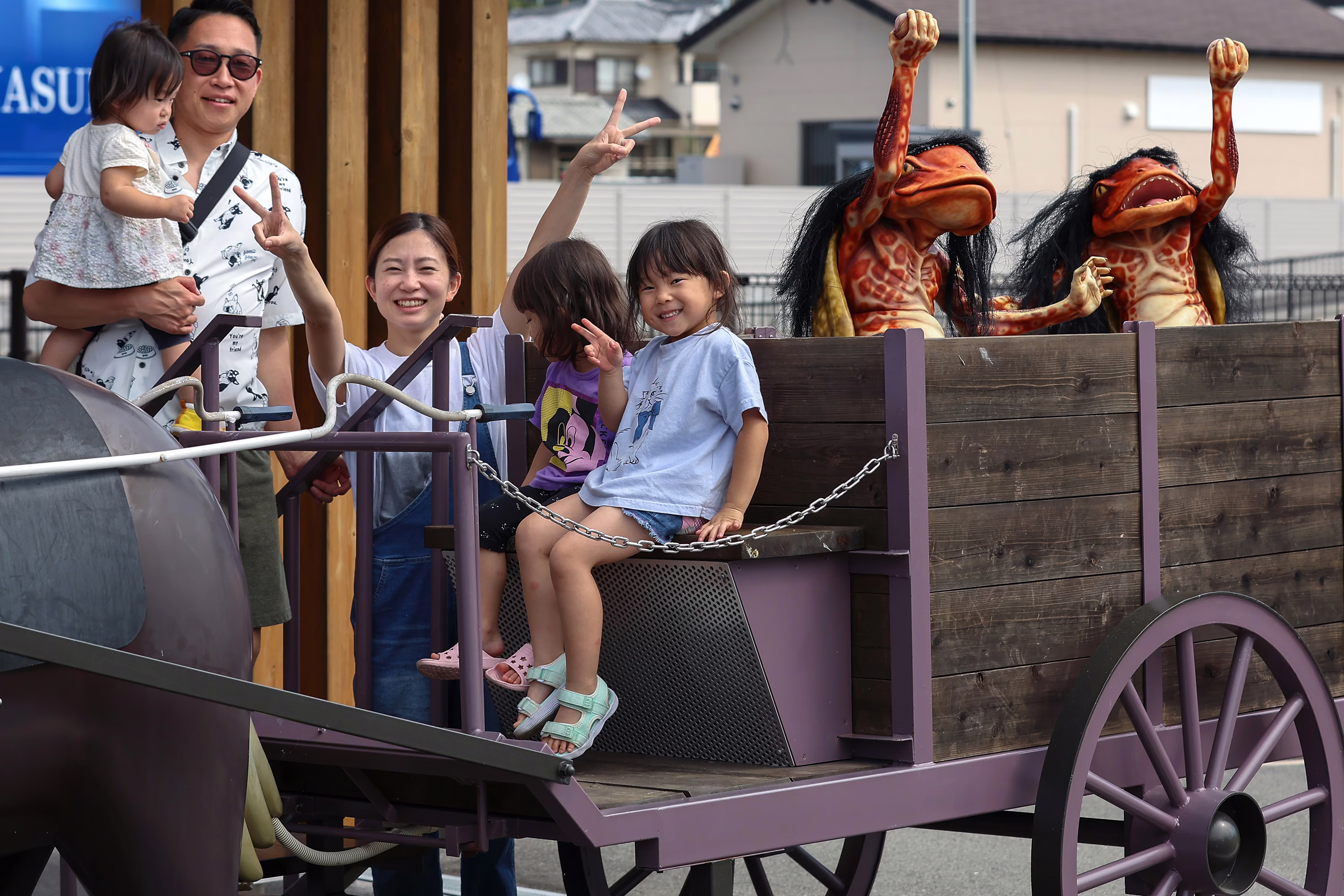 A family with young children poses for a photograph on a wagon, beside statues of two frightening-looking monsters in the back of the wagon.