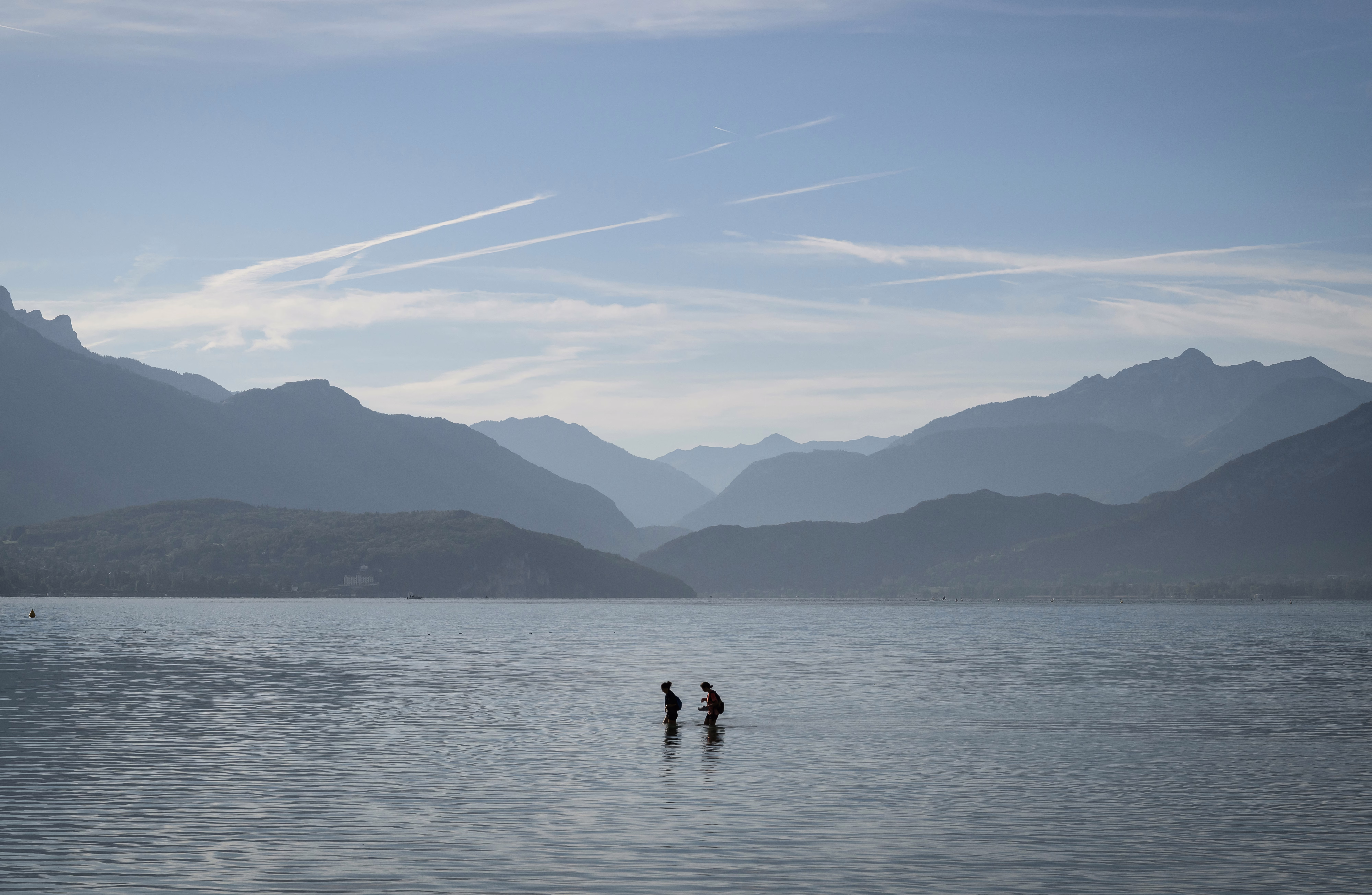 A wide view of two people wading through shallow water in a broad lake, with mountains visible in the background