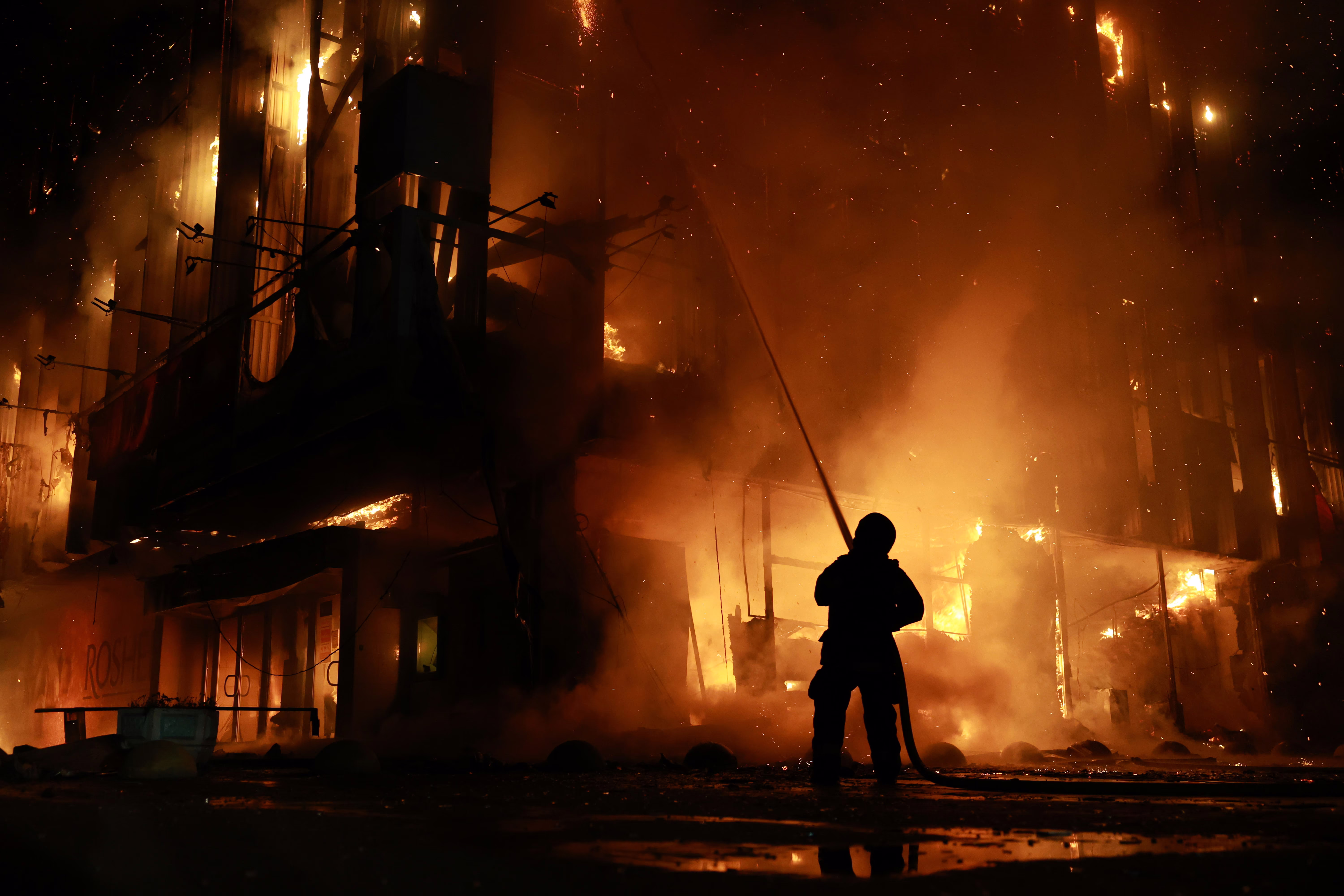 A person uses a hose to spray water onto a tall burning building at night.