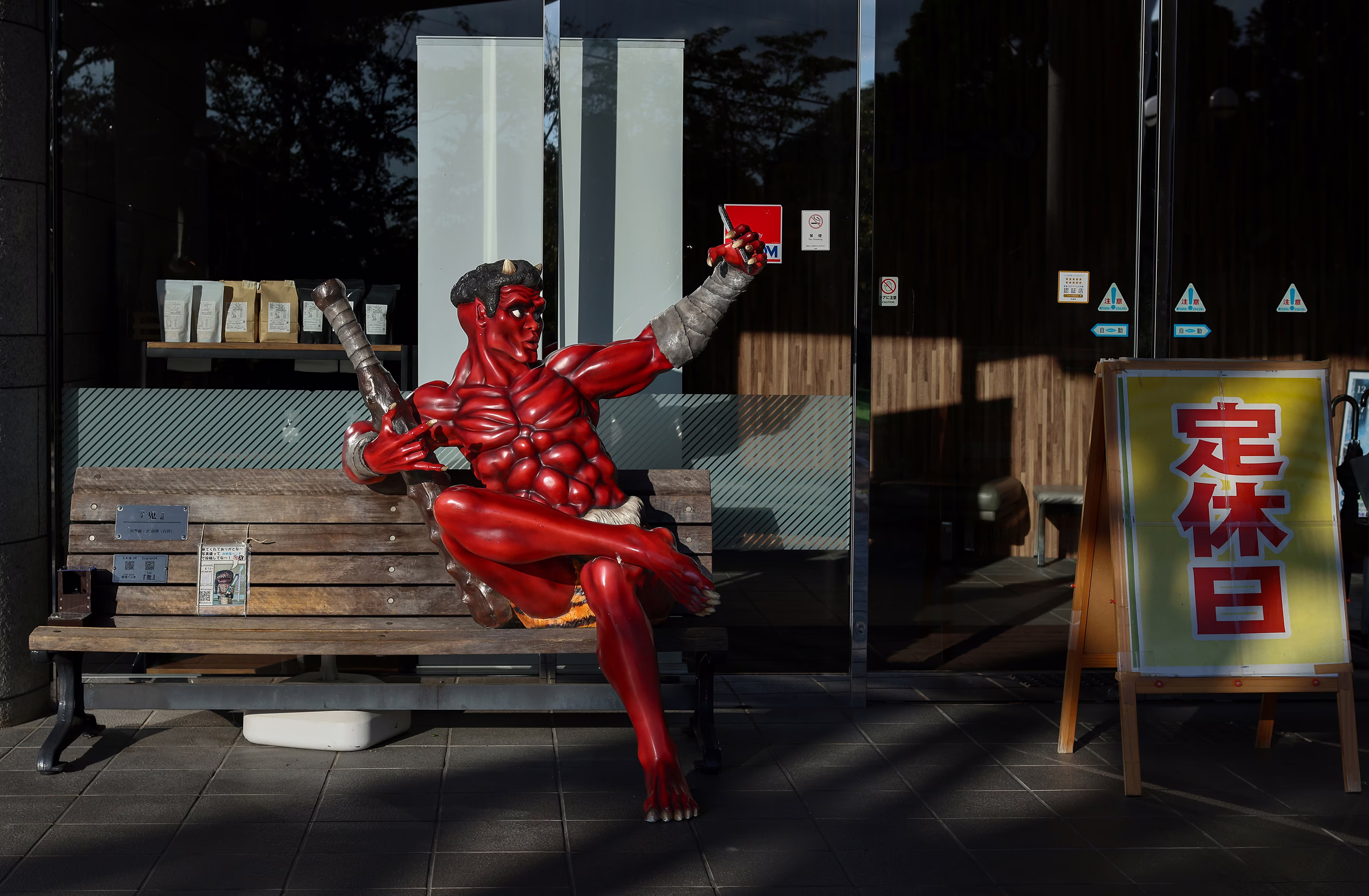 A statue of a demon taking a selfie sits on a bench in front of a storefront.