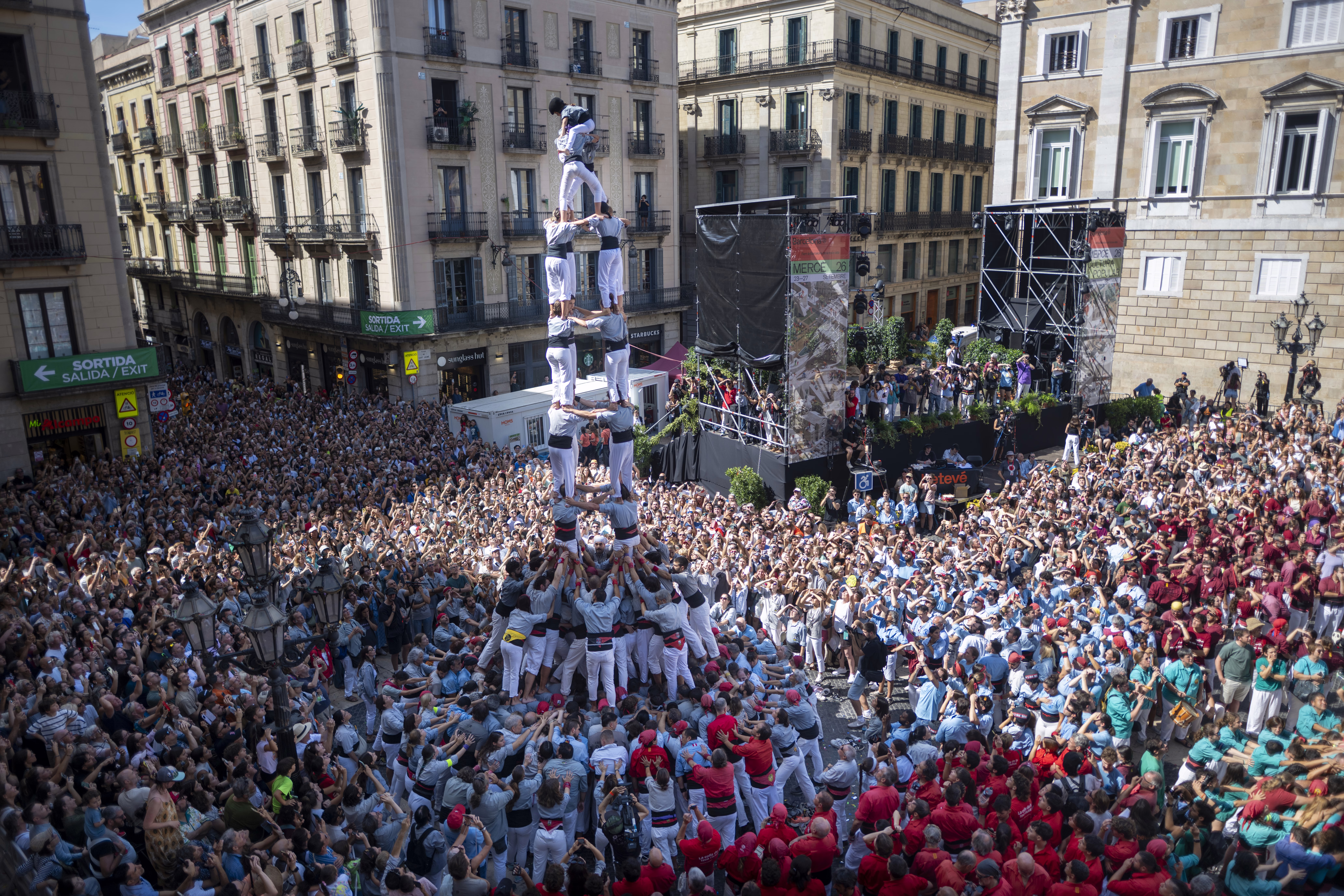 A large crowd of people packs into a square in a town, watching a group that is forming a tall human tower.