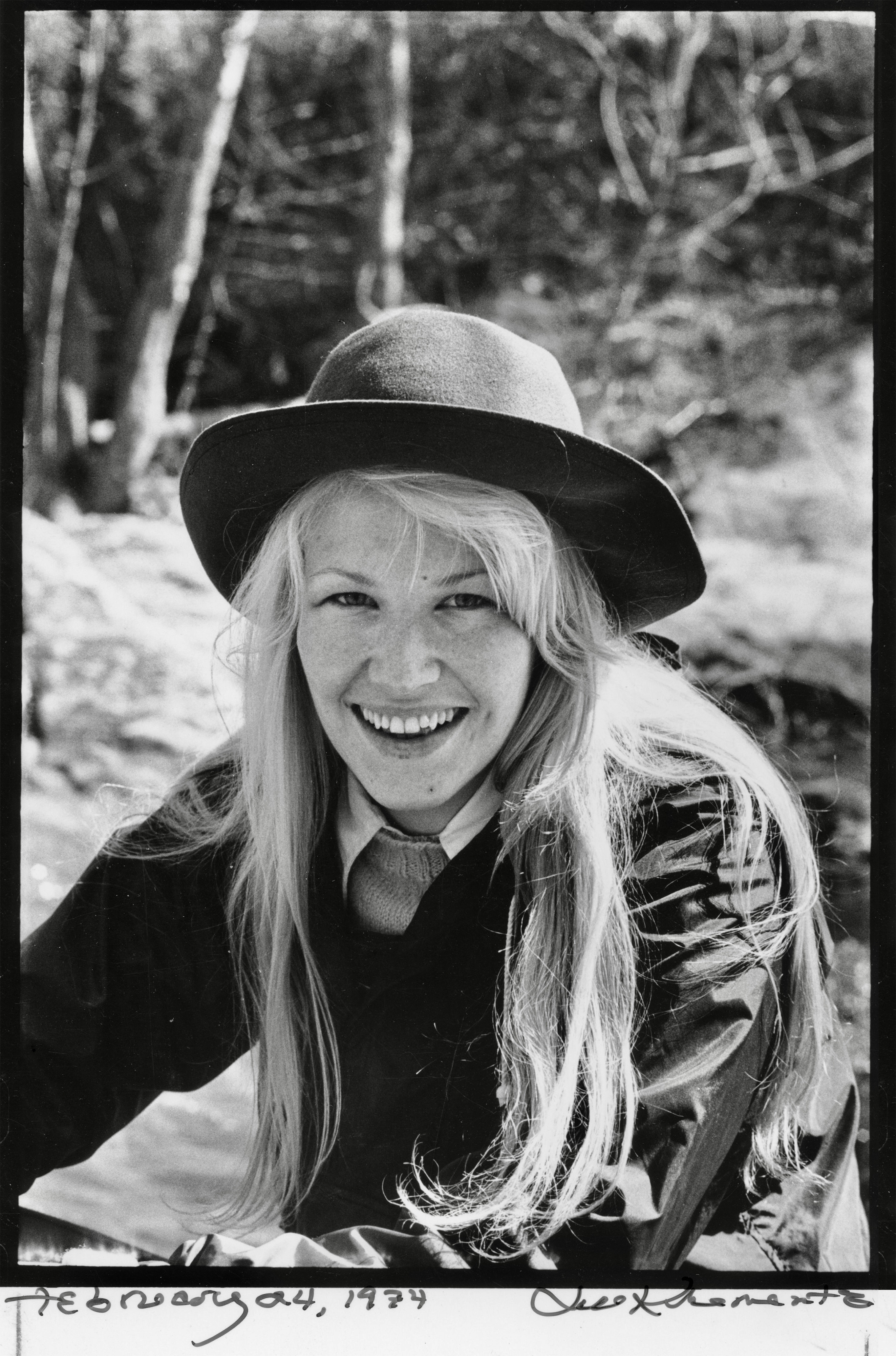 A black-and-white photograph of Annie Dillard, smiling against a backdrop of trees while wearing a round felt hat