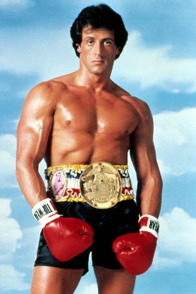 Sylvester Stallone as Rocky Balboa, bare-chested and wearing black boxing shorts, red boxing gloves, and a gold championship belt.