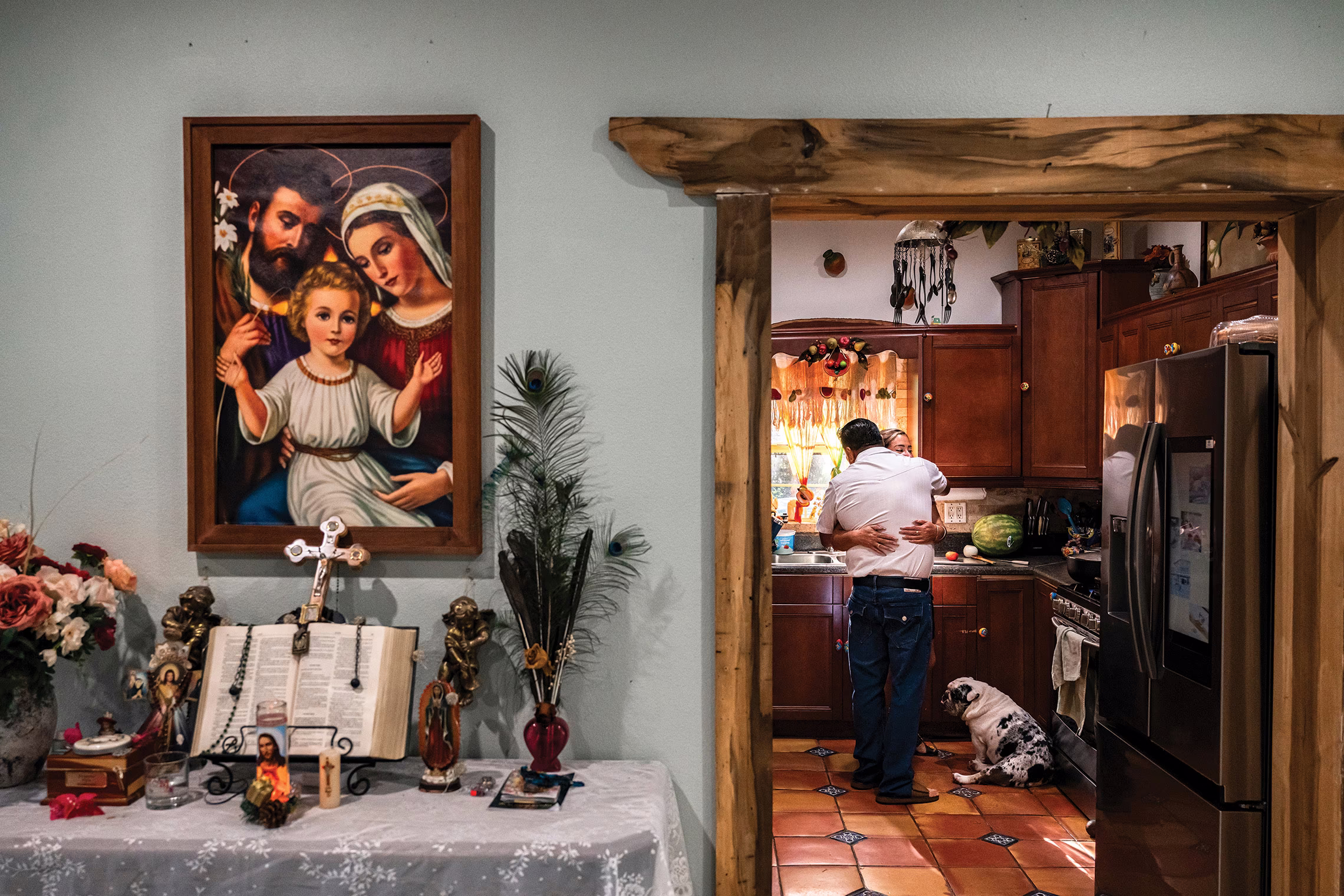 photo of room with painting of the holy family, side table with open bible and cross, and doorway looking into tiled kitchen, where two people embrace and a dog looks on