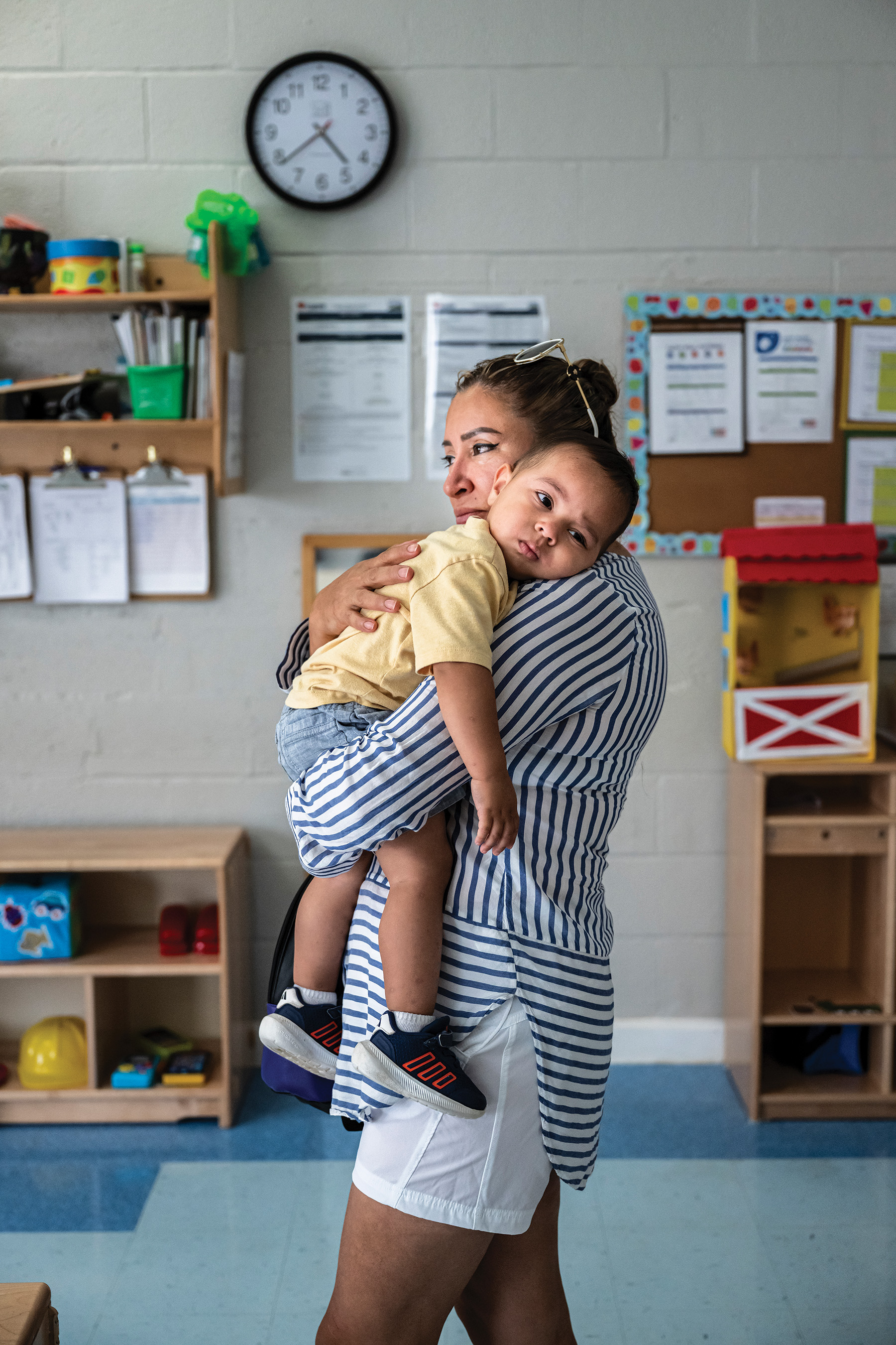 photo of woman in striped shirt and white shorts standing in children's classroom and holding toddler with his head on her shoulder