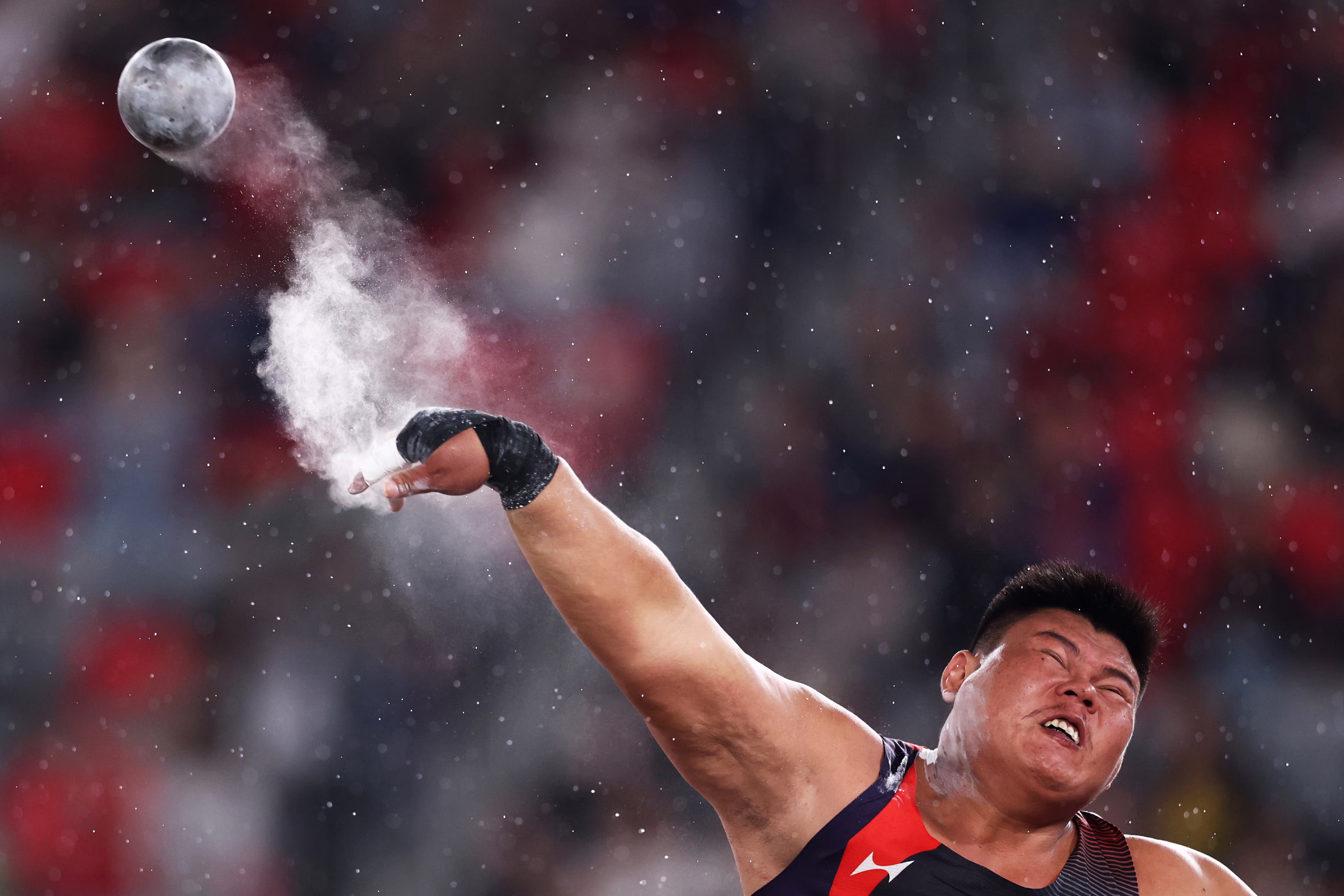 An athlete releases a shot put, a small cloud of chalk coming off their hand.