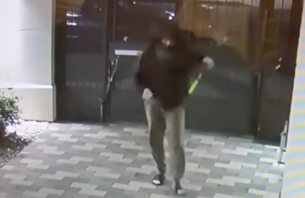 Surveillance footage of a man in dark clothing and light pants climbing over a gate.