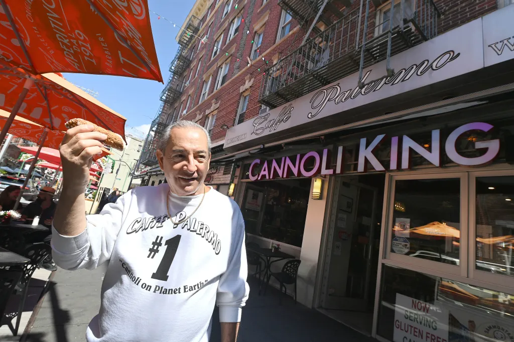 John DeLutro, wearing a white sweatshirt, holds up a cannoli outside Caffe Palermo on Mulberry Street.