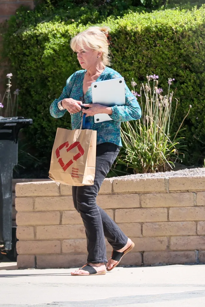Linda Purl walking outdoors with a shopping bag and a laptop.