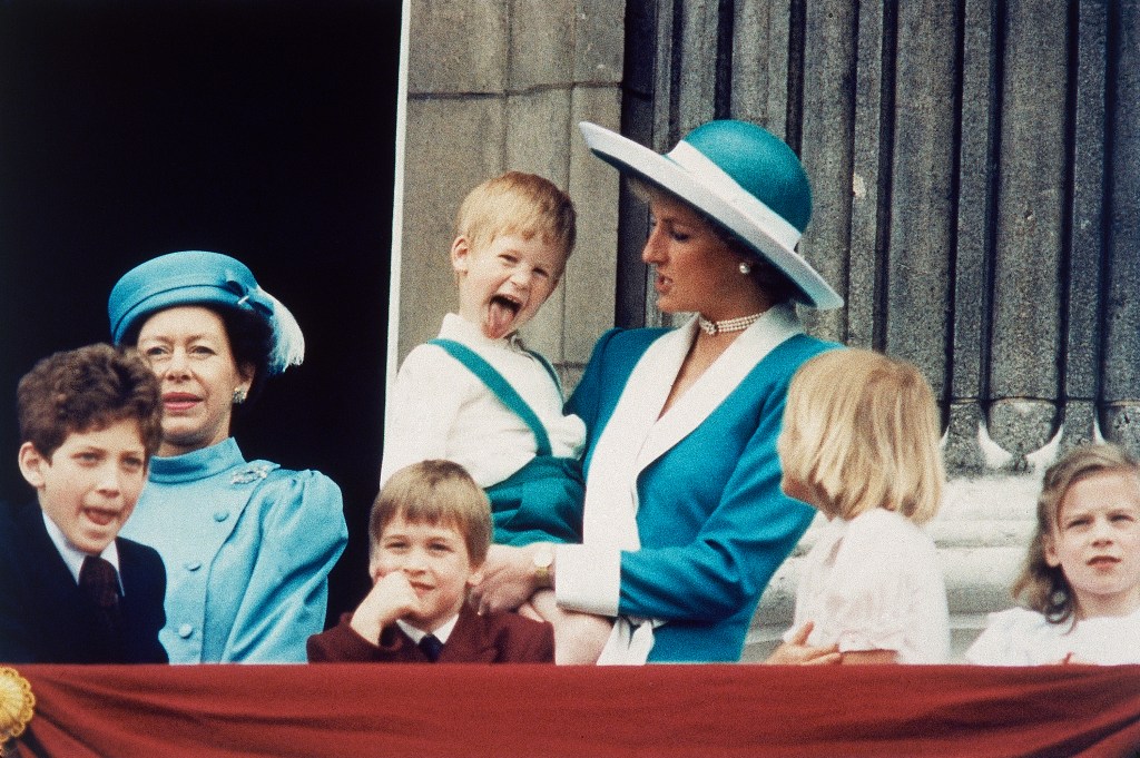 Princess Diana, in a turquoise blue coat with white trim and matching hat, holding young Prince Harry, in matching turquoise overalls and a white shirt, who sticks out his tongue, on the balcony of Buckingham Palace.