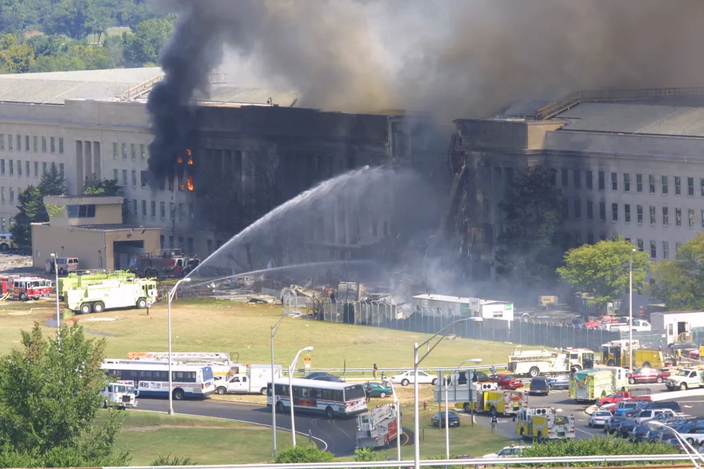 Smoke and flames rise from the west wing of the Pentagon building after a plane crash, with firefighters spraying water and emergency vehicles gathered on the surrounding grounds.