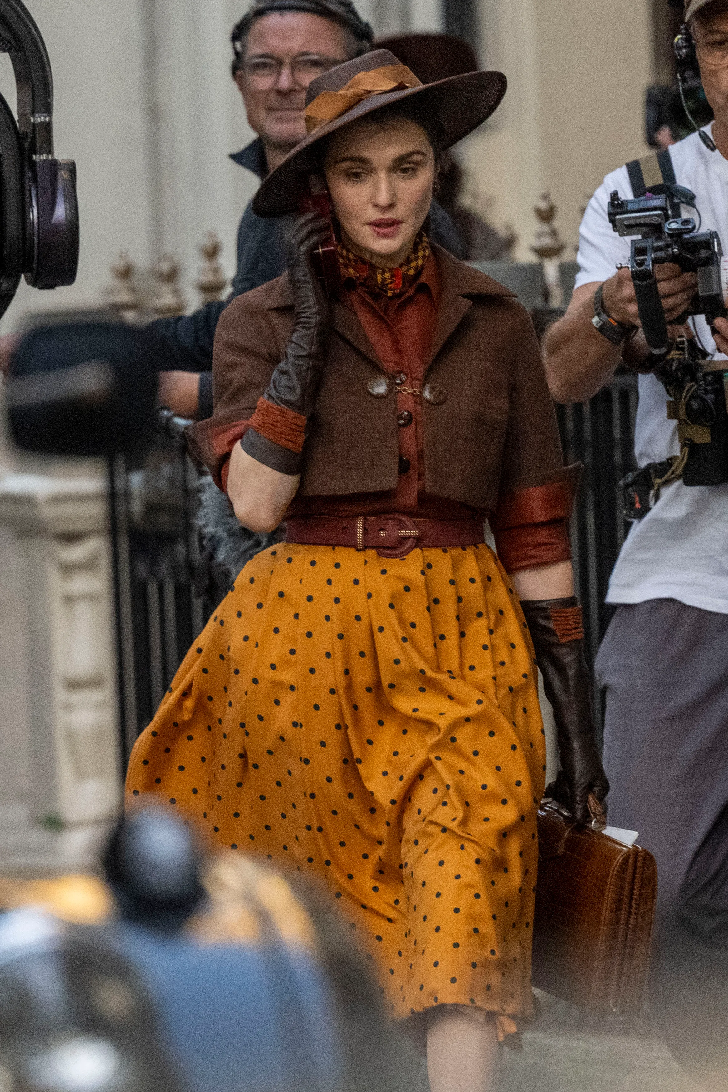Rachel Weisz as Evelyn O'Connell in a mustard polka-dot skirt, brown cropped jacket, and wide-brimmed hat, holding an old-fashioned transmitter radio to her face on the set of The Mummy.
