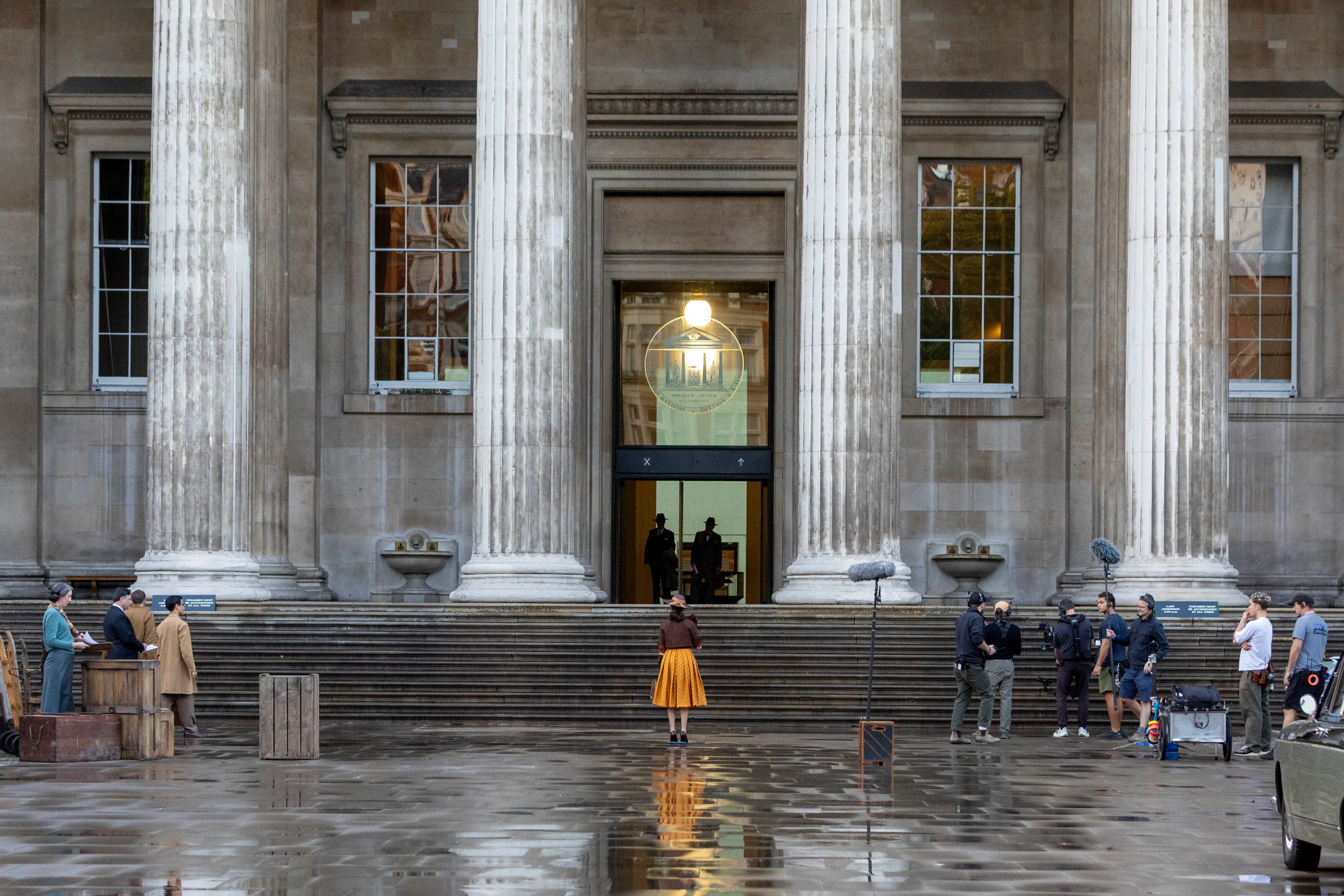 Rachel Weisz as Evelyn O'Connell on the set of The Mummy, wearing a mustard yellow polka-dot skirt, brown cropped jacket, and matching wide-brim hat, standing in front of a grand building with large columns.
