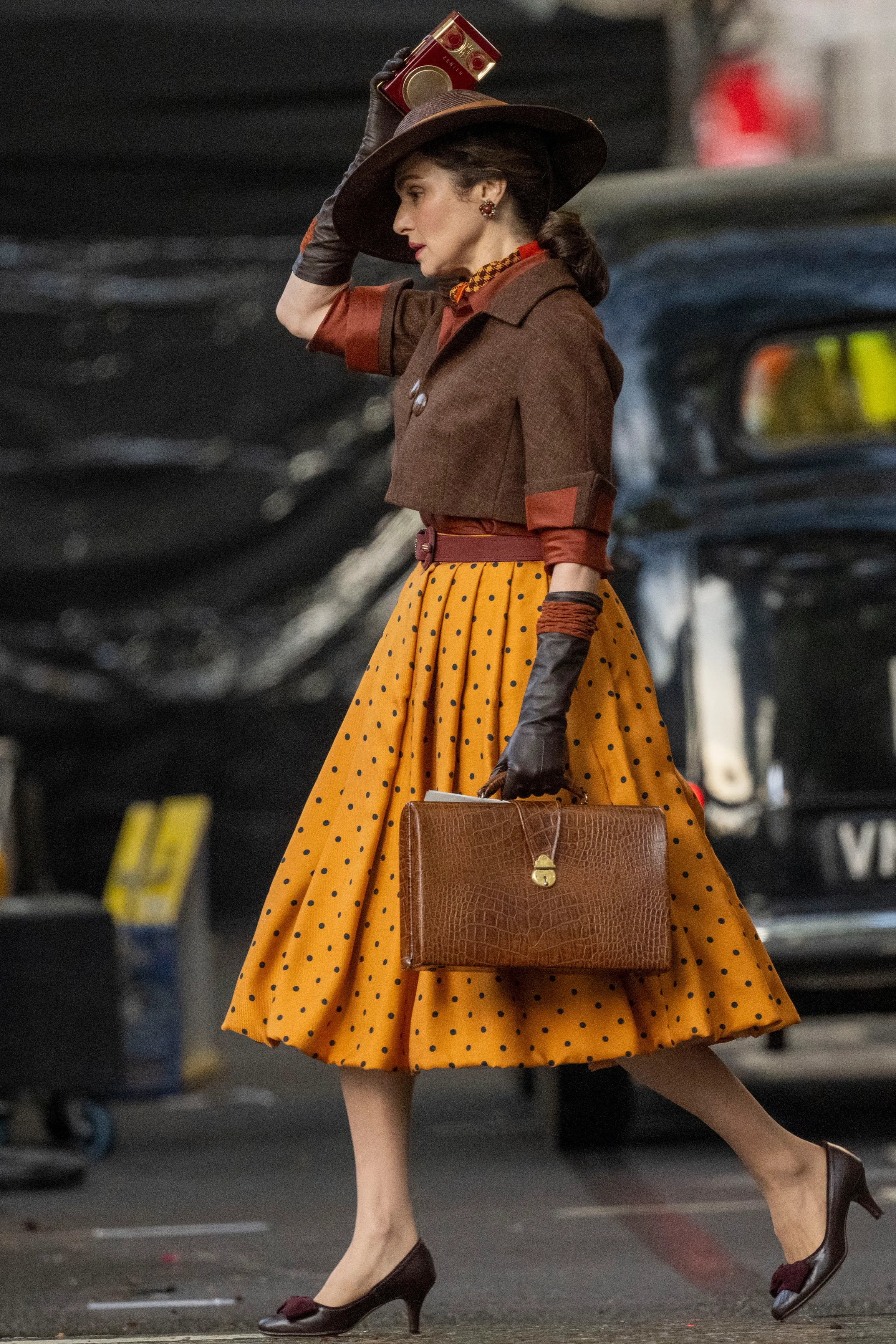 Rachel Weisz as Evelyn O'Connell in a yellow polka dot dress, brown jacket, and hat, holding a radio to her ear and carrying a briefcase.