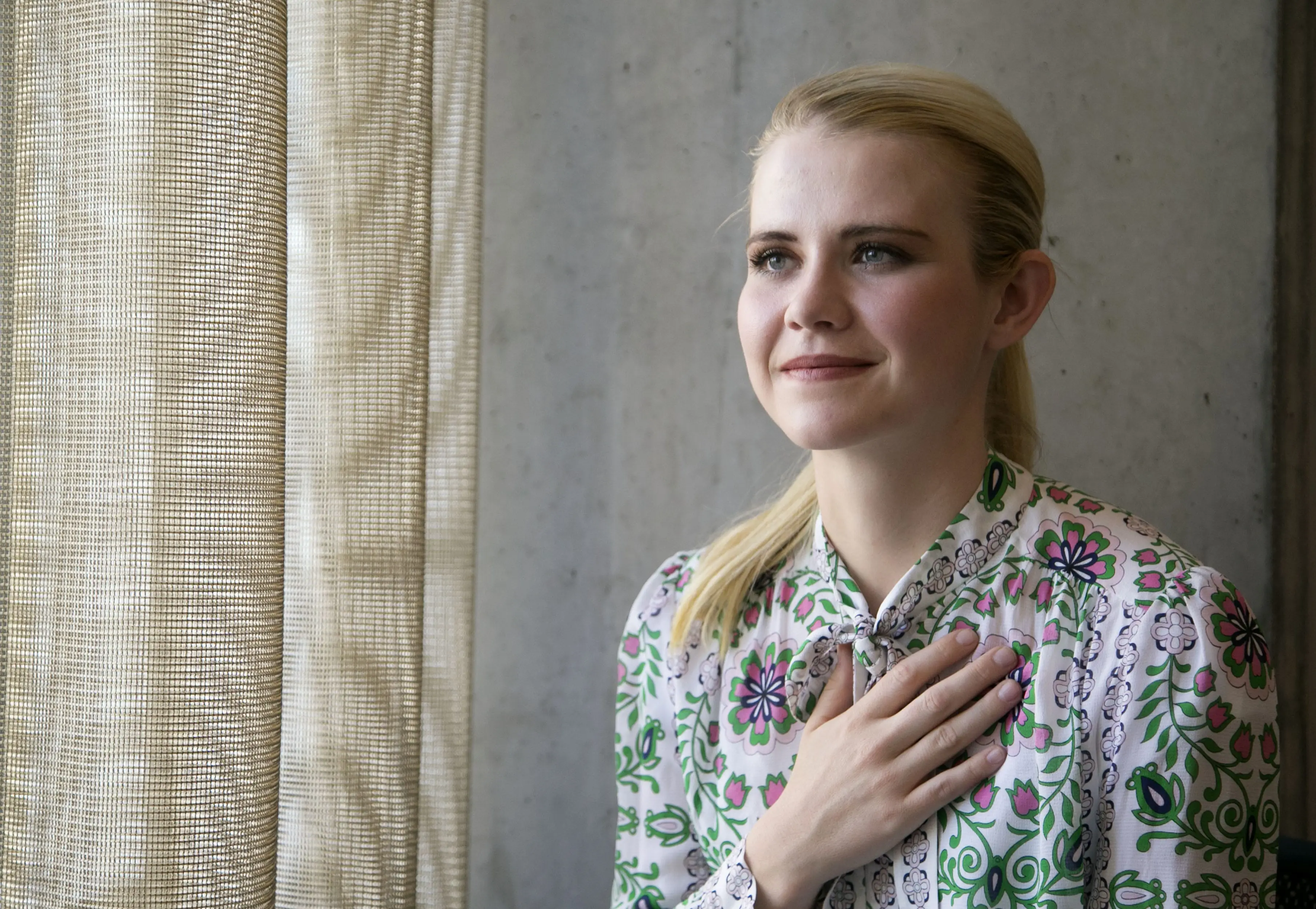 Elizabeth Smart, a woman with blonde hair, wearing a white blouse with green and pink floral patterns, looks out a window with her hand over her heart.