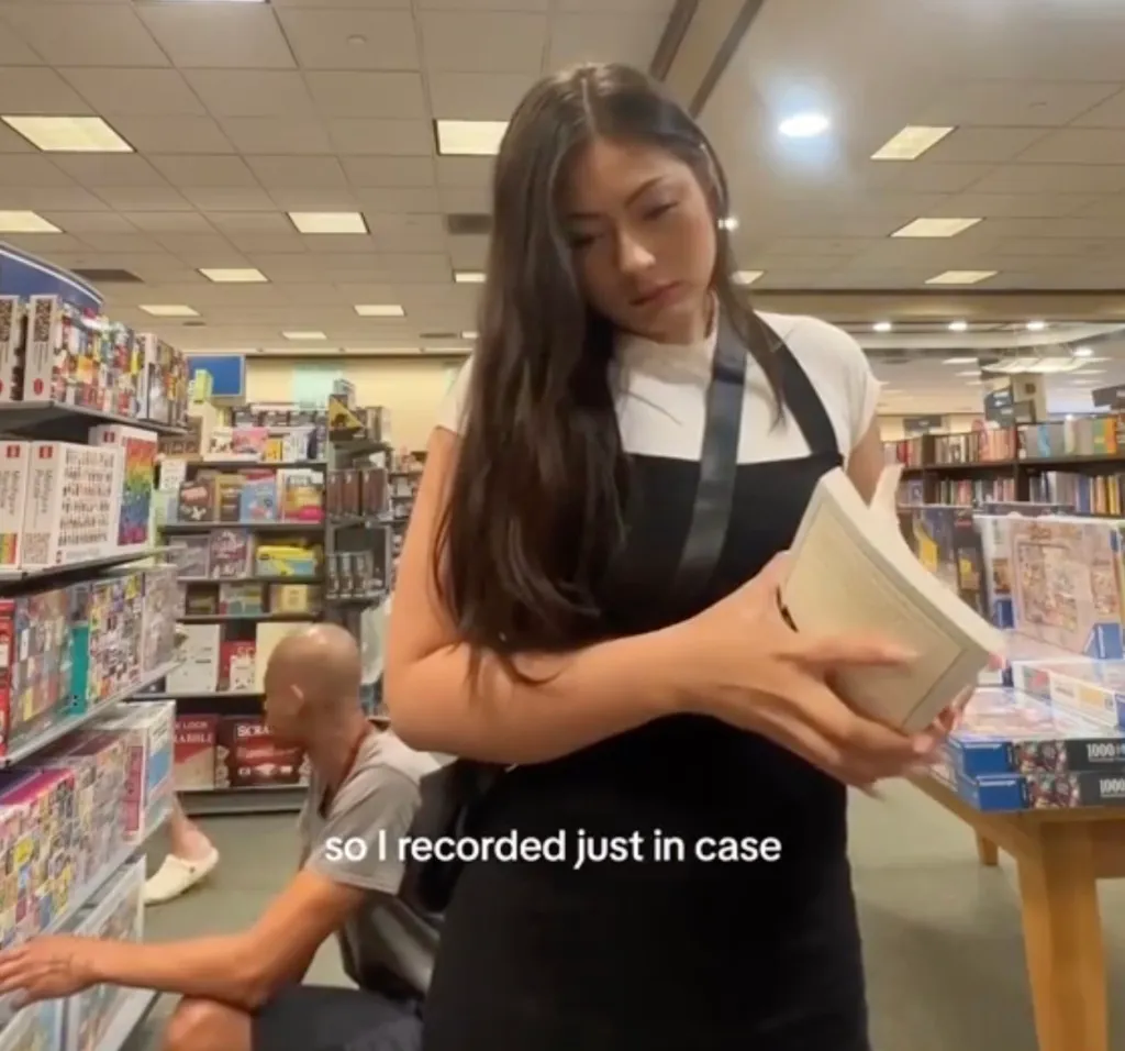 A woman in a bookstore holding a book, with a man crouching behind her.